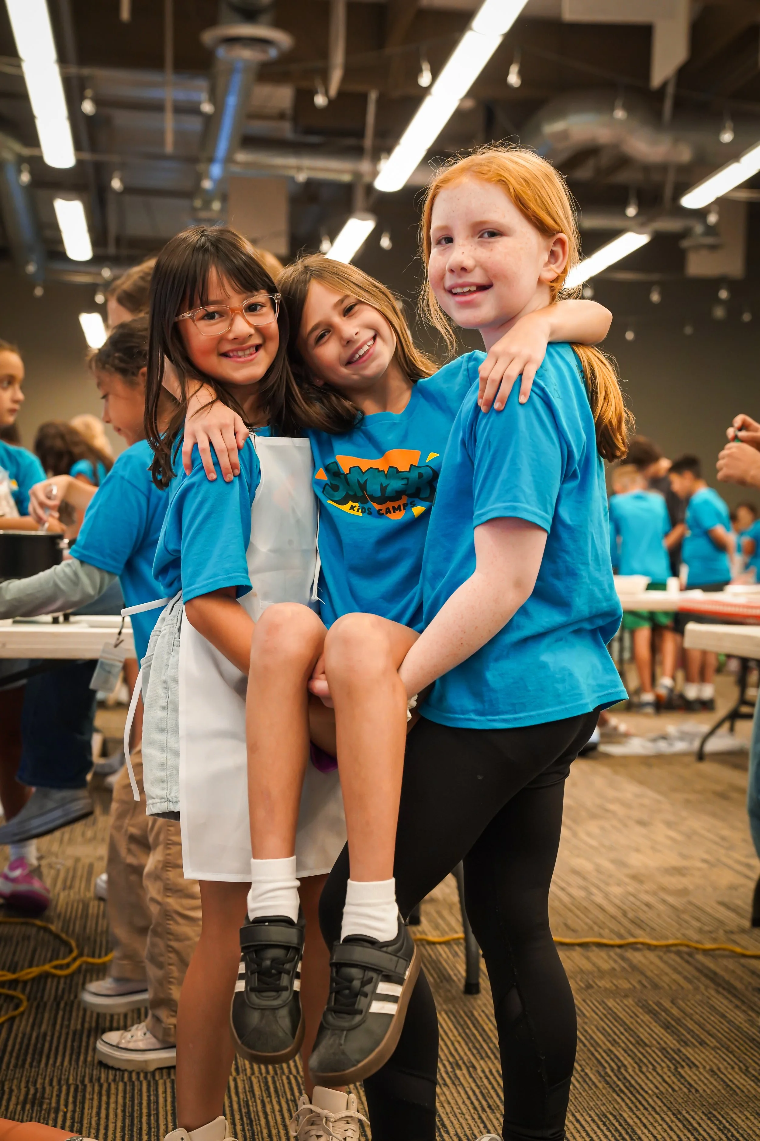 Group of children at a summer camp, with a girl being lifted by two others, all smiling and wearing blue camp t-shirts.