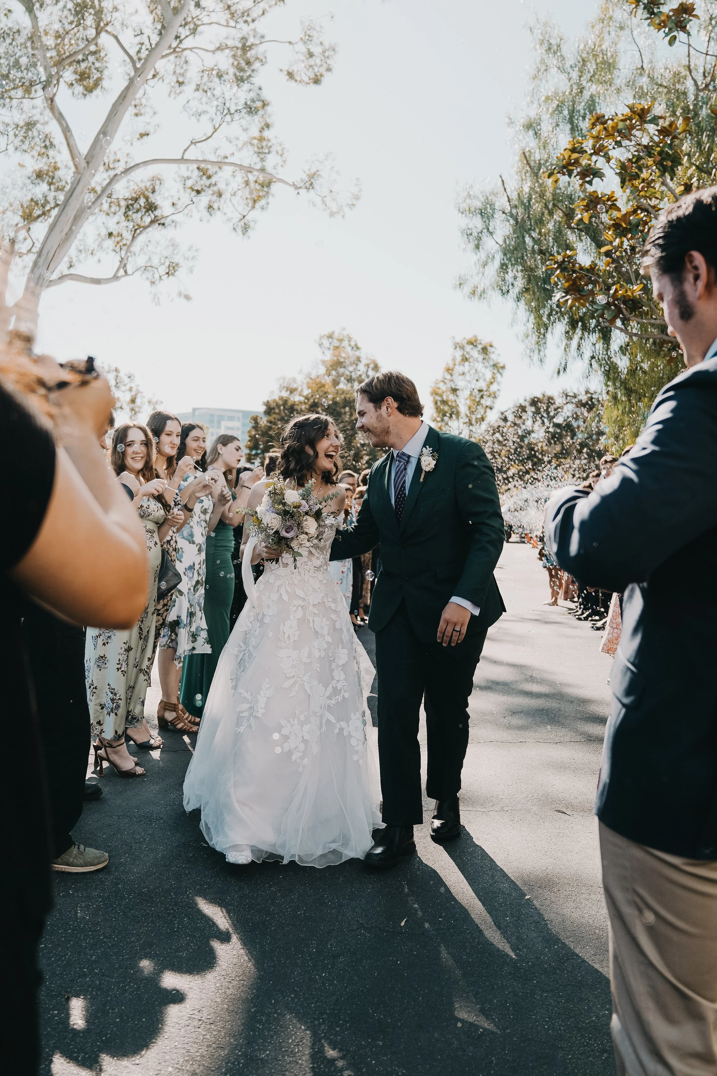 Bride and groom smiling and holding hands during their outdoor wedding celebration, surrounded by cheering guests.