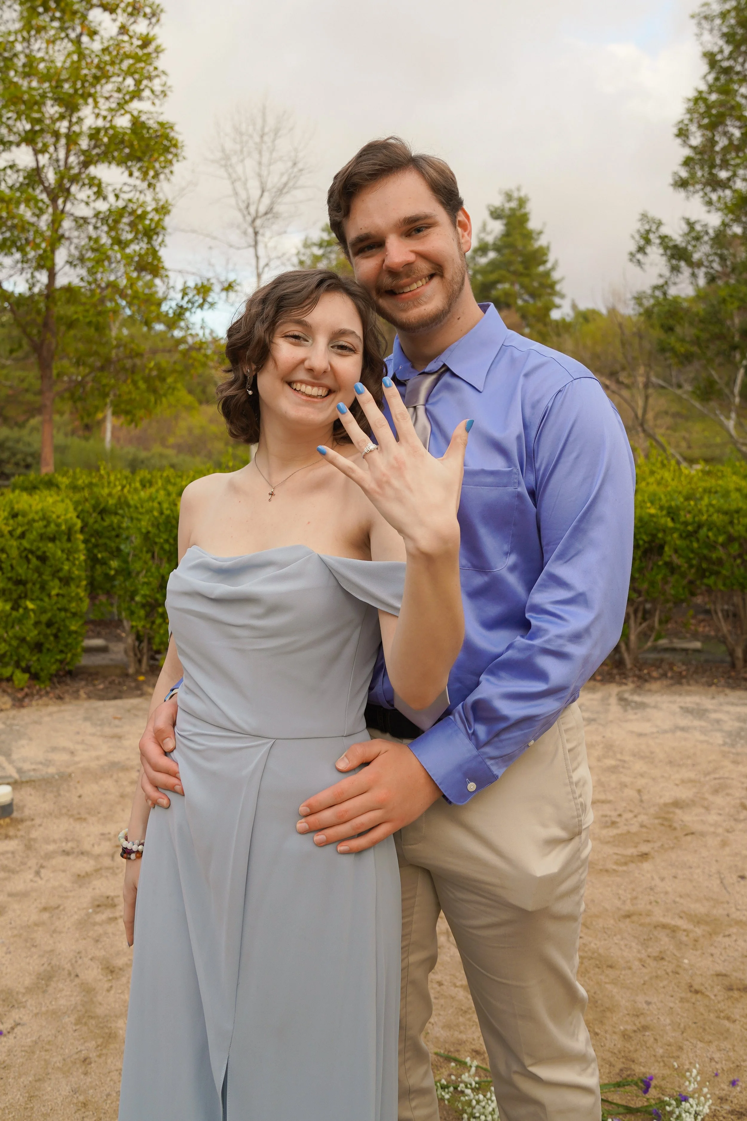 A young couple stands outdoors, smiling. The woman is displaying her engagement ring and has her nails painted blue. She is wearing a strapless gray dress, and he is wearing a light blue dress shirt with khaki pants.