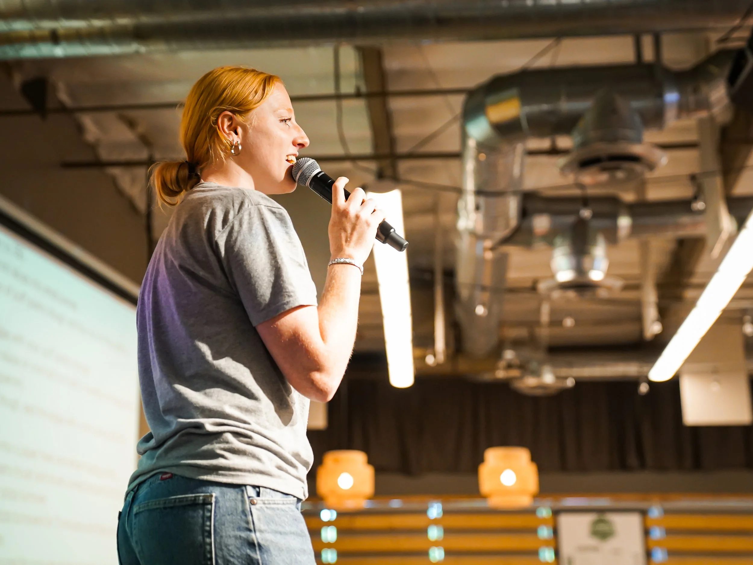 A woman with red hair in a ponytail speaking into a microphone during a presentation in a modern indoor setting.