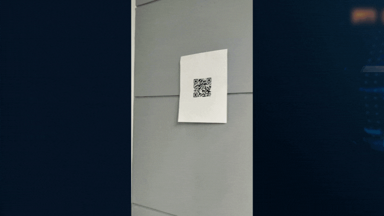 A QR code printed on a white piece of paper taped to a gray tiled wall.