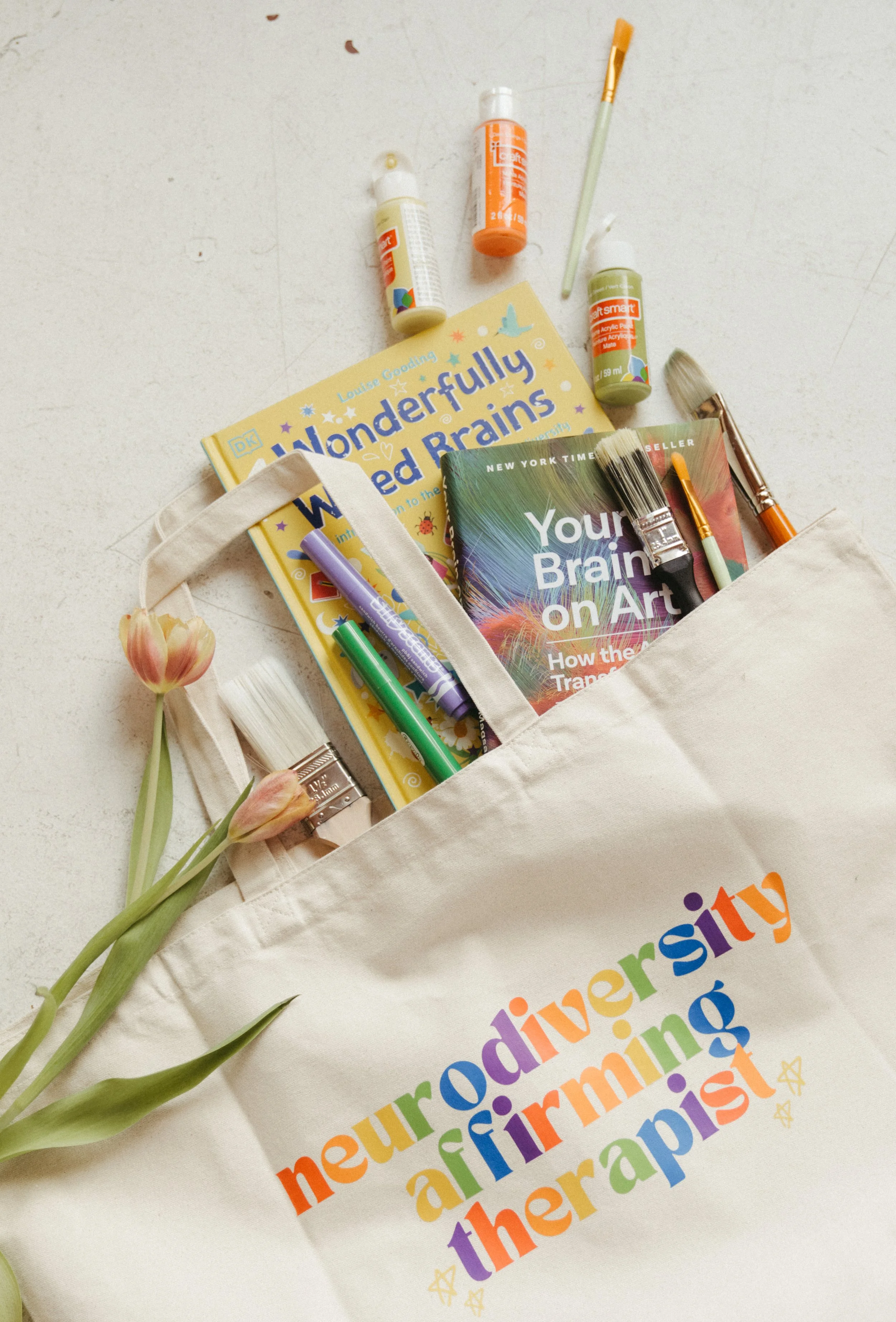 Canvas tote with art supplies and books. Bag reads in rainbow letters "neurodivergent affirming care"