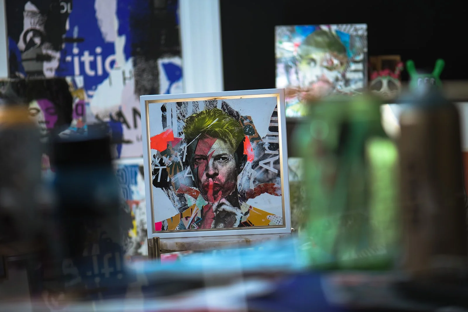 Vibrant artwork featuring a mixed media portrait in a studio setting, with blurred spray paint cans in the foreground and colorful, abstract art in the background.