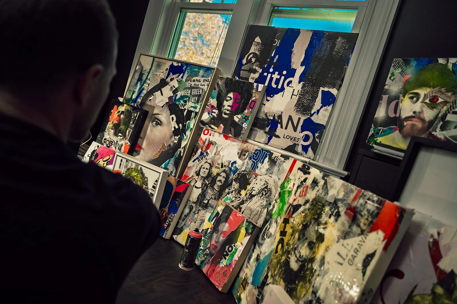 Various colorful abstract and mixed media artworks leaning against a wall in a studio setting, with a person observing them. The artworks feature collage-style portraits and vibrant colors.