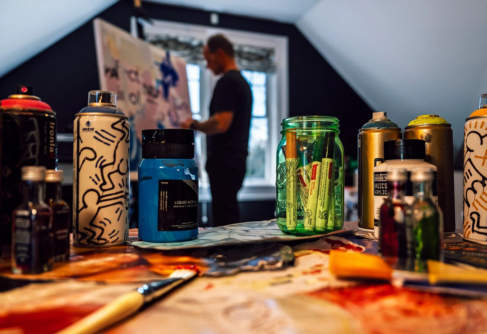 Artist's studio with spray paint cans and acrylic paints on a table, artist painting in the background.