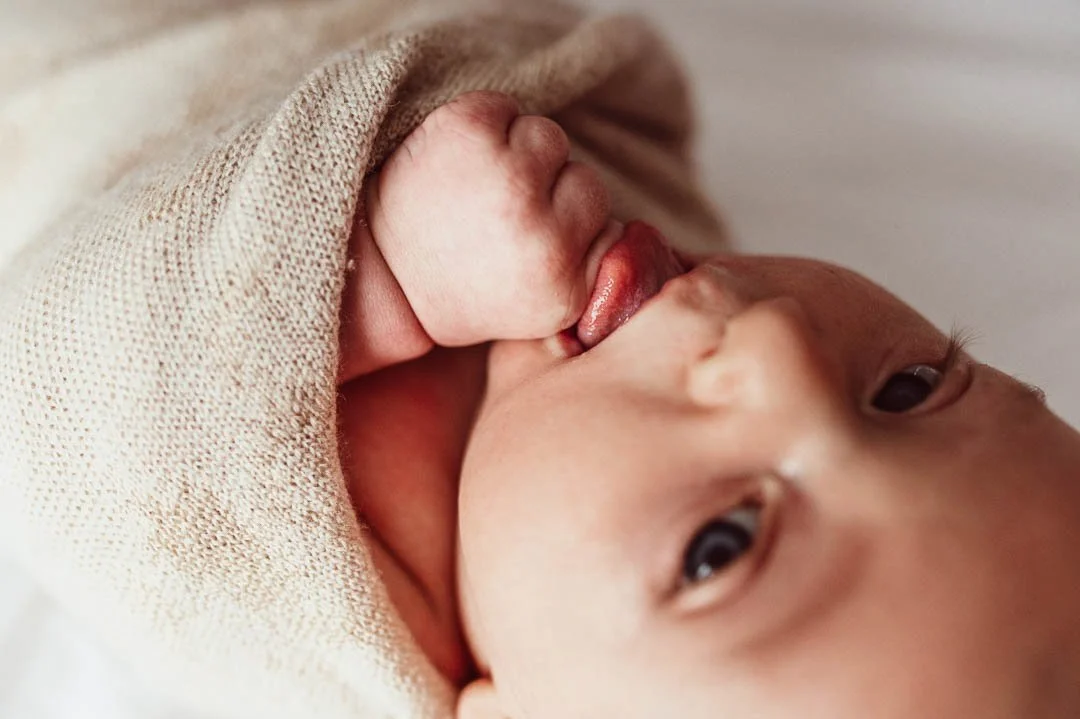 A baby swaddled in a neutral-toned wrap, sucking on his hand in a relaxed moment. during newborn session