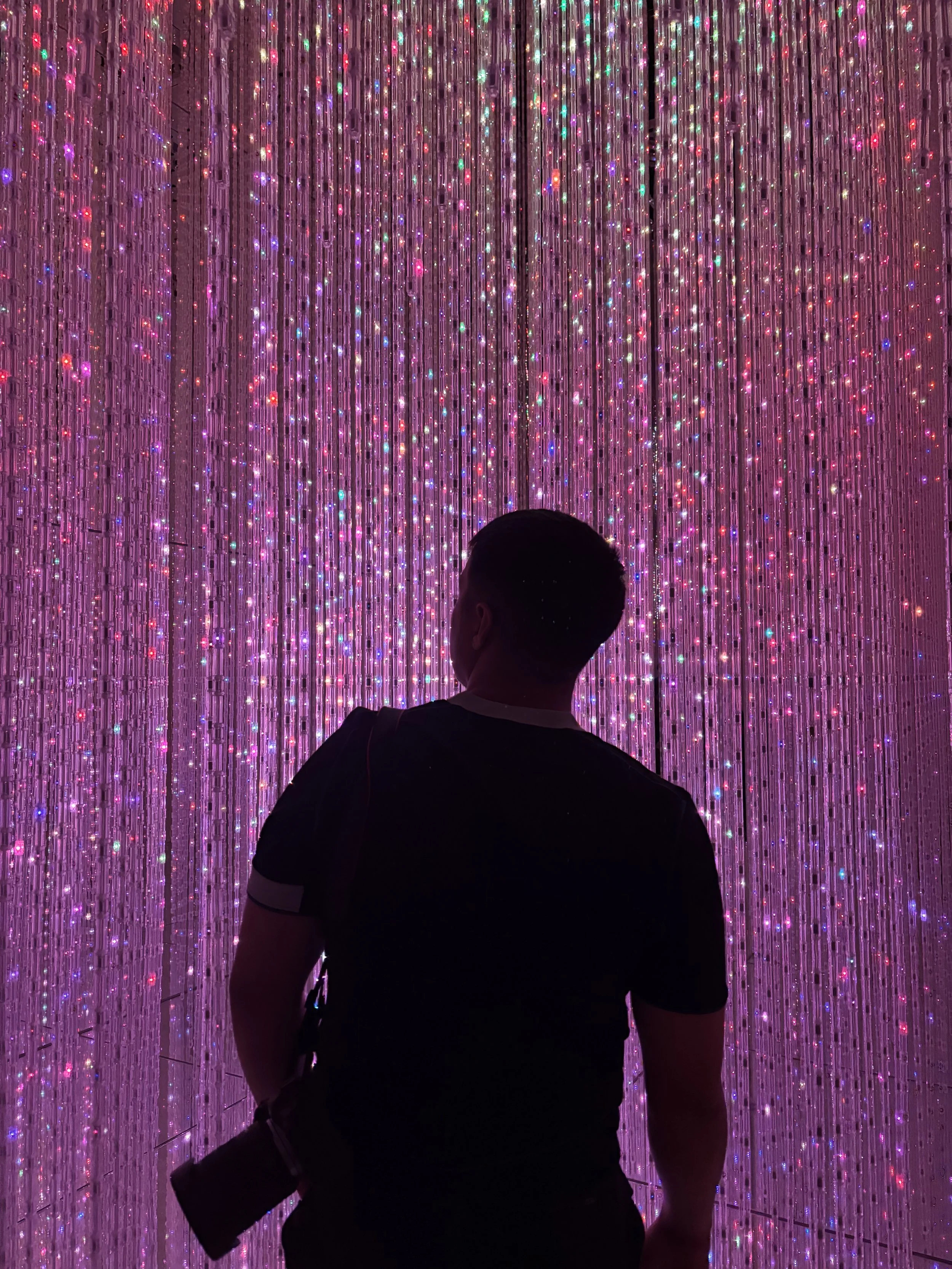 Person standing in front of a colorful LED light installation with purple, pink, and multicolored lights reflecting off hanging strings.