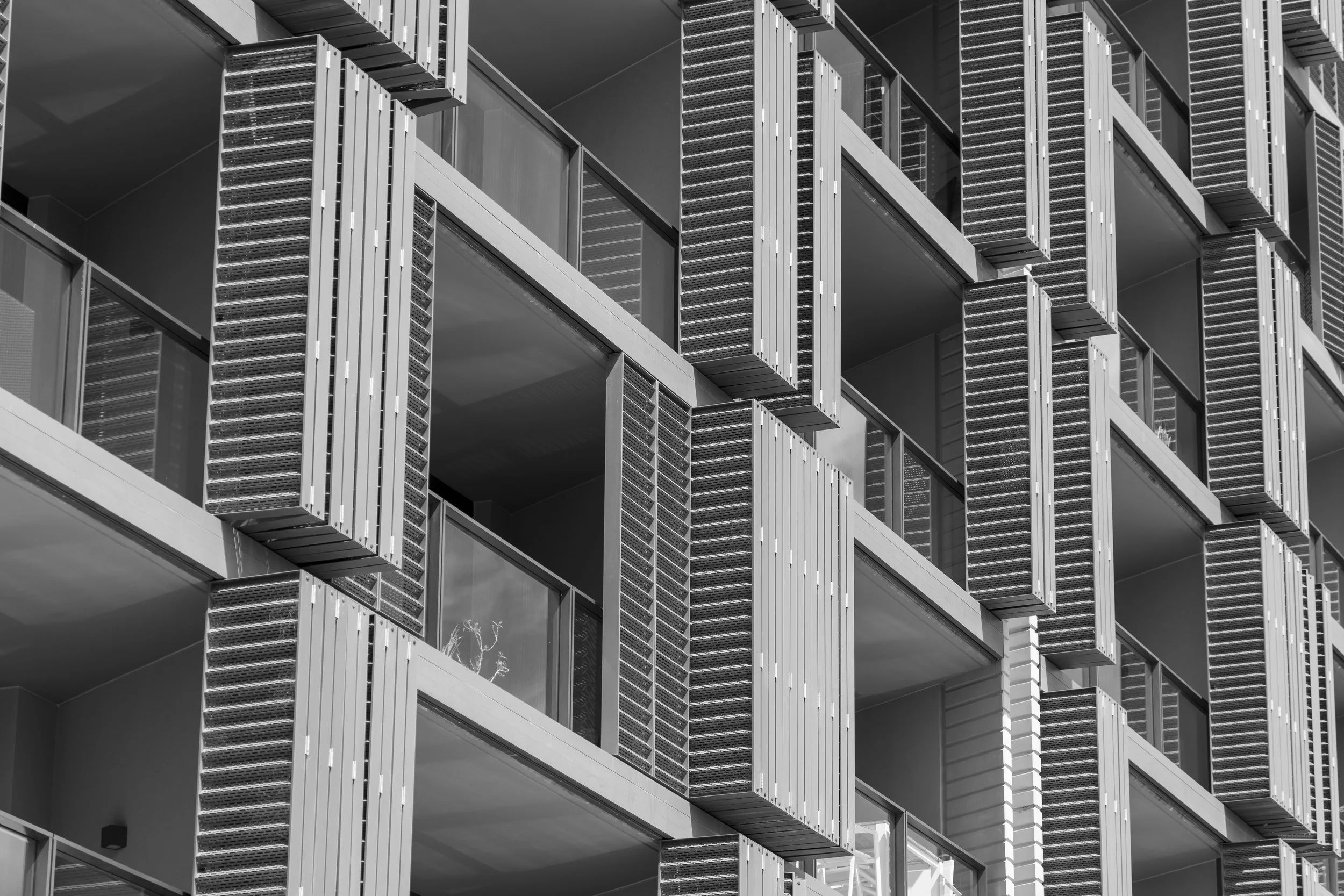 Black and white photo of a modern apartment building with multiple balconies, each featuring window shutters or louvered panels.