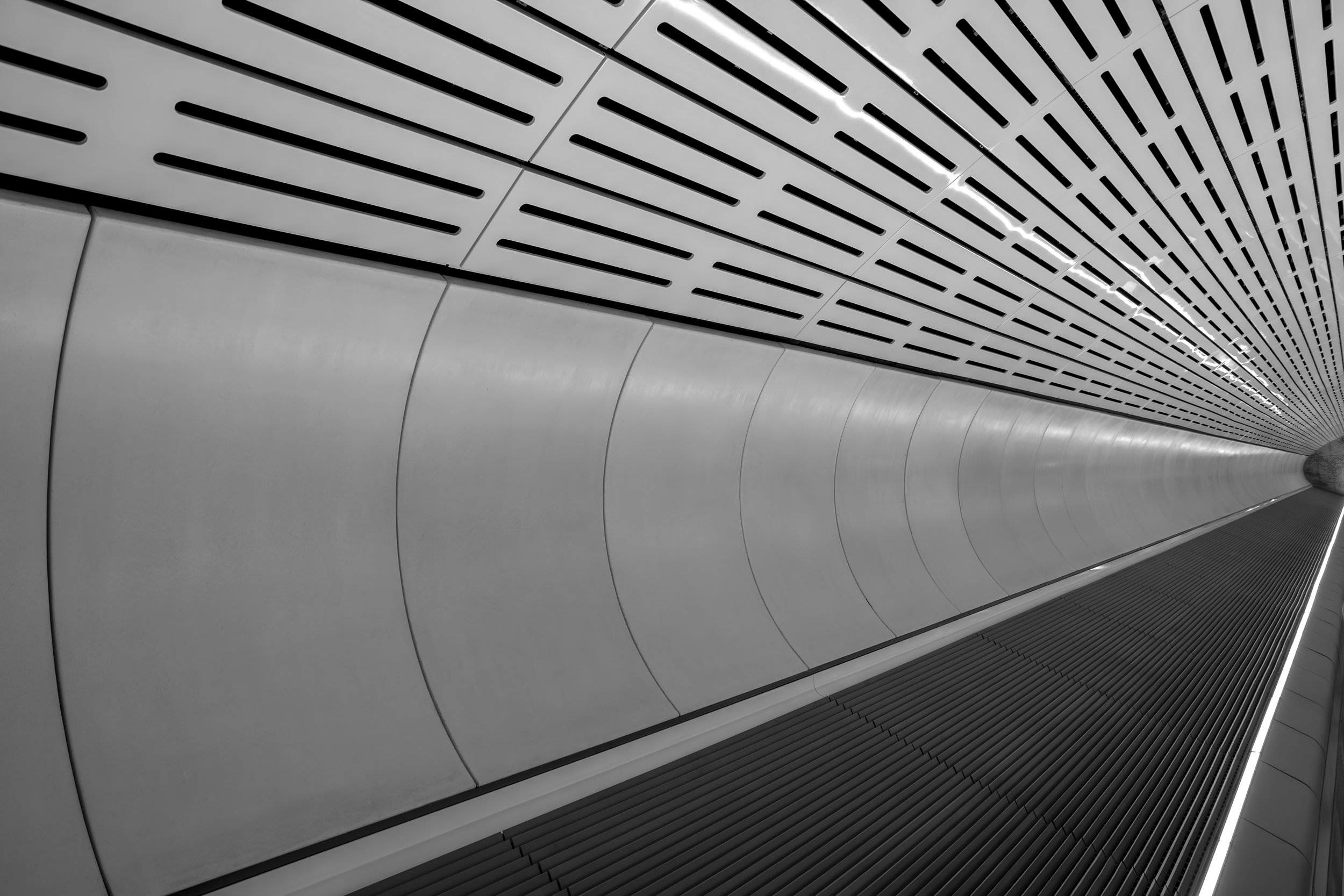 Looking up at a modern, futuristic tunnel with smooth, curved gray walls and a perforated metal ceiling with linear vents.
