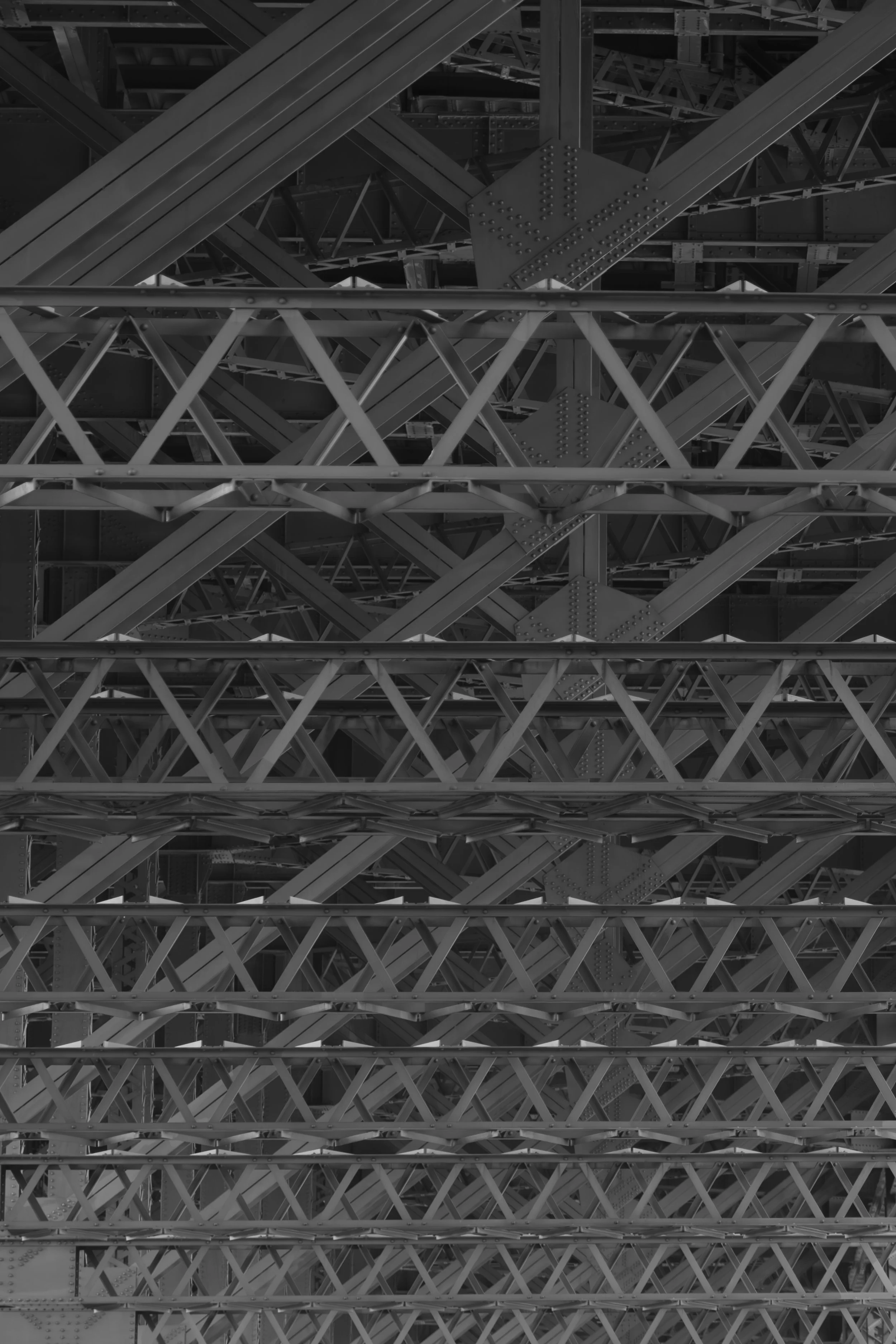 Black and white photograph of the underside of a bridge showing a complex network of steel girders, beams, and rivets.