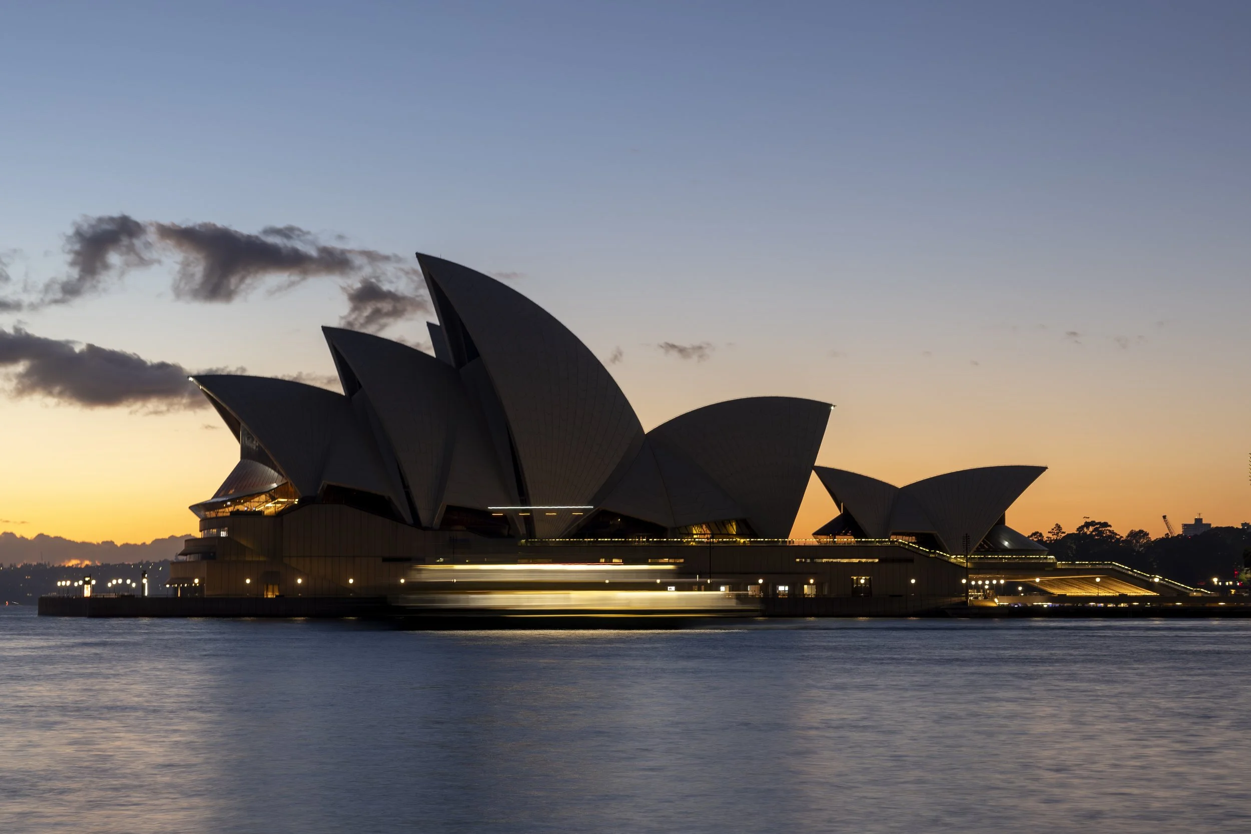 Sydney Opera House at sunset with clouds in the sky and water in the foreground.
