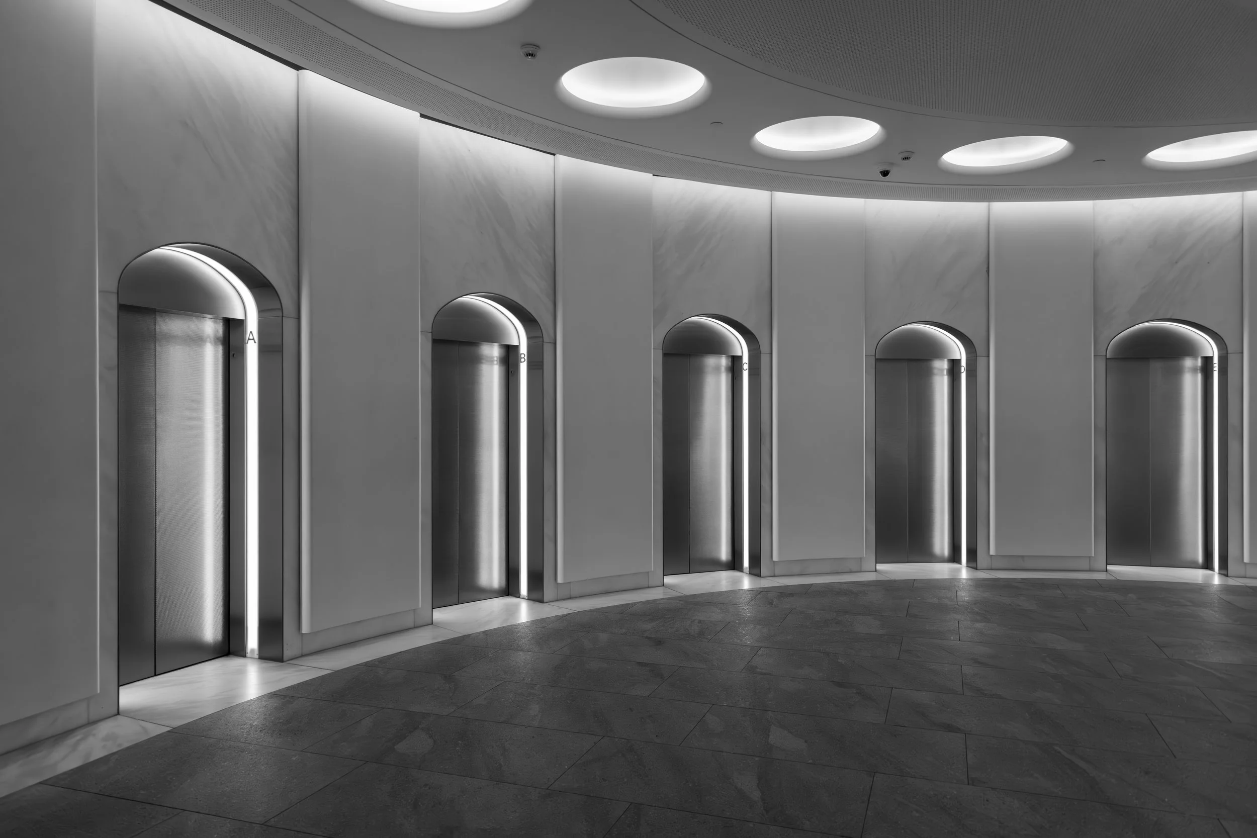 A modern, sleek elevator bank with five stainless steel elevator doors and arched trim, set in a minimalist, gray-toned lobby with a textured floor and circular ceiling lights.