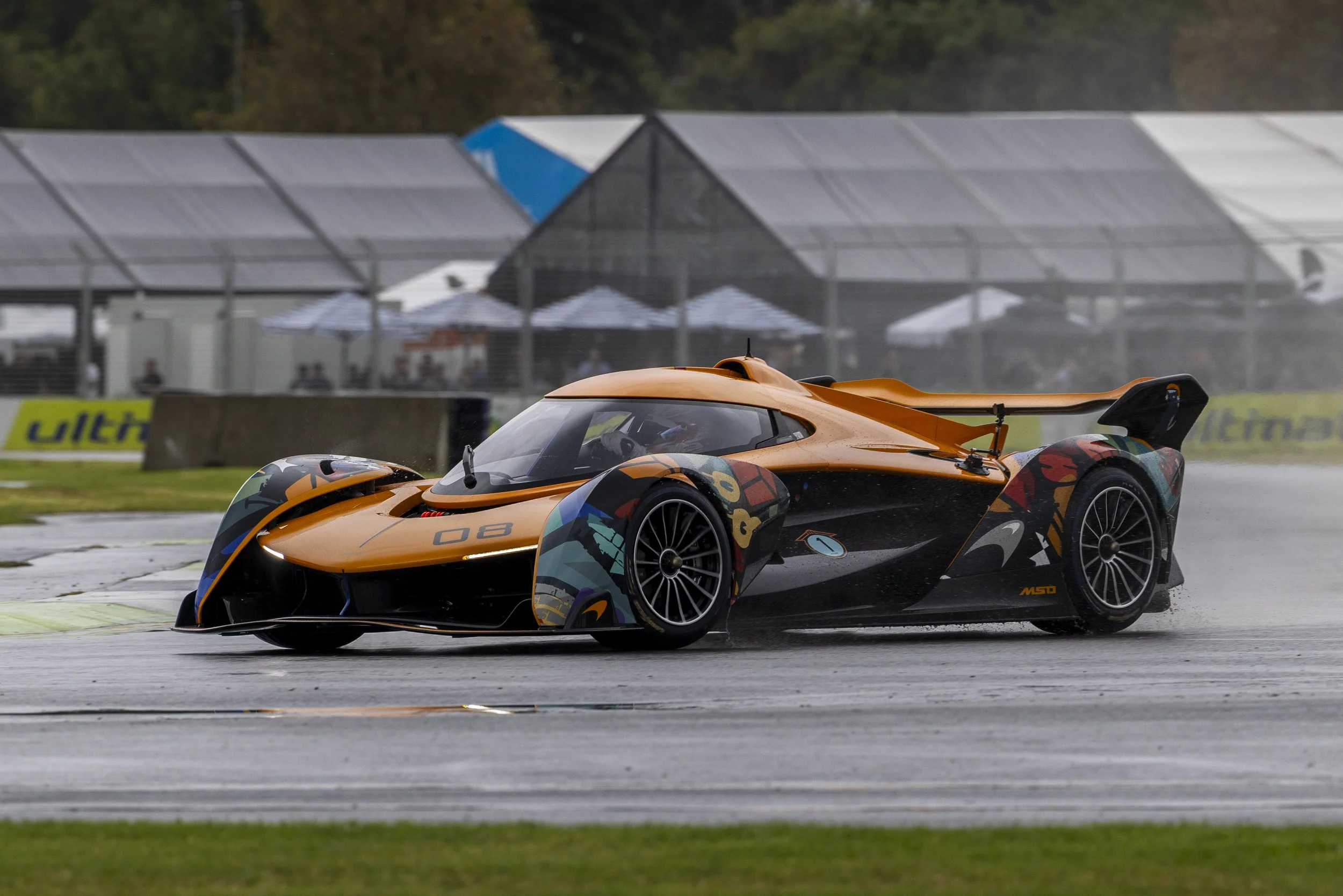 An orange race car with a colorful abstract design racing on a wet track during rain with blurred spectators and tents in the background.