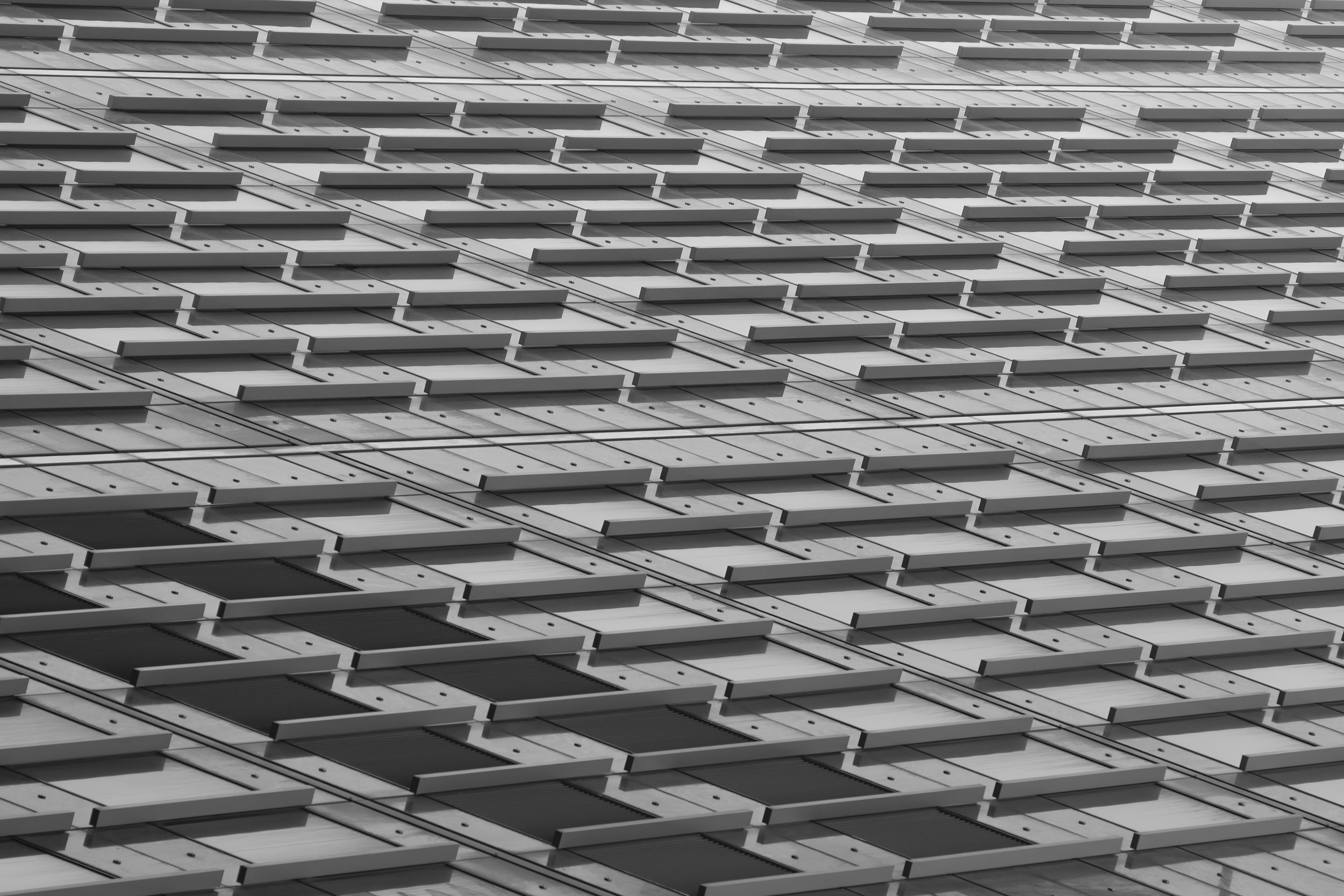 A black-and-white photograph of a modern building's exterior, showing a pattern of rectangular windows and external panels arranged in a repetitive and geometric design.