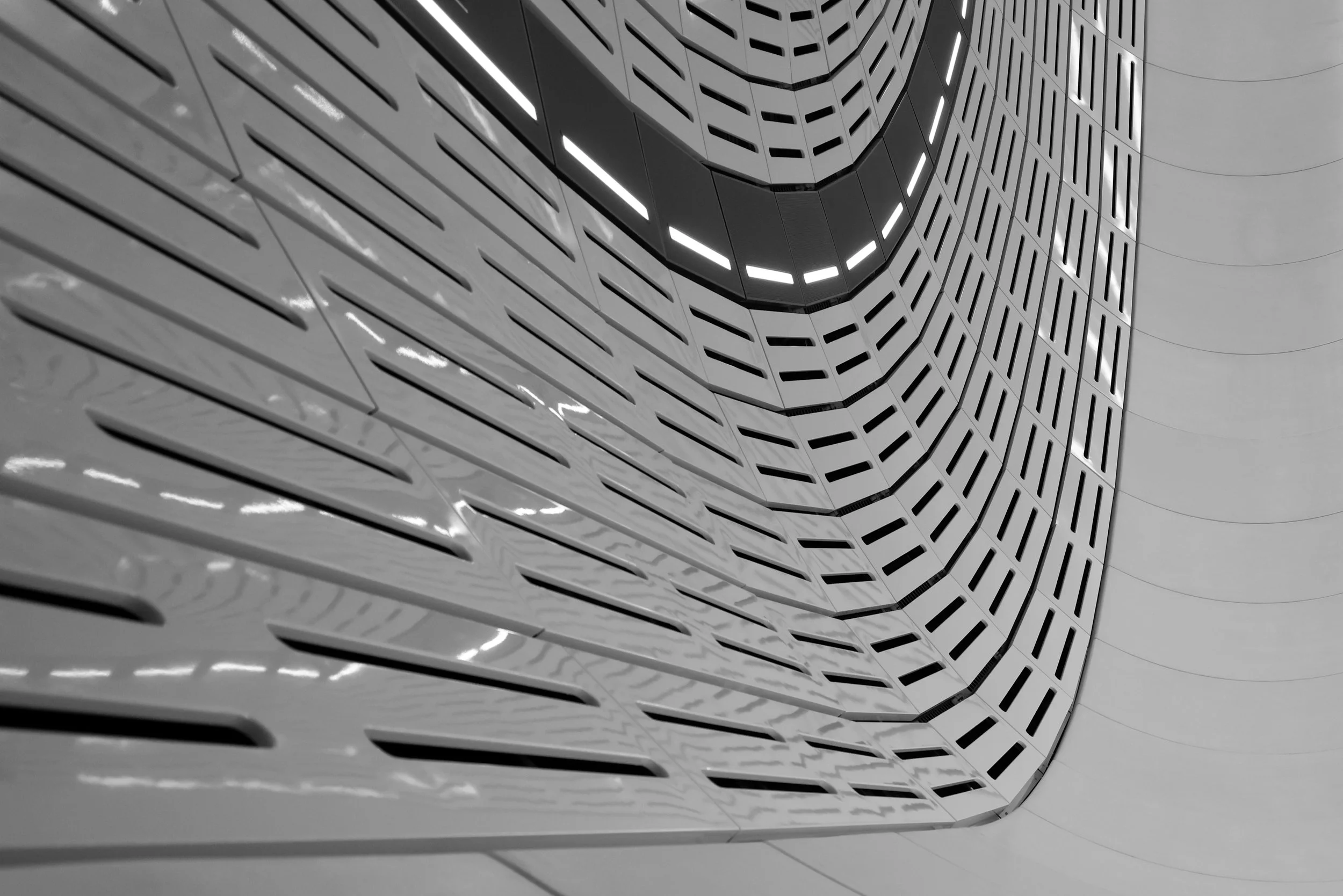 A modern architectural interior with a curved wall featuring horizontal, elongated, rectangular cutouts. The structure has a sleek, futuristic design with black and white tones, and illuminated lines accentuate its curved shape.