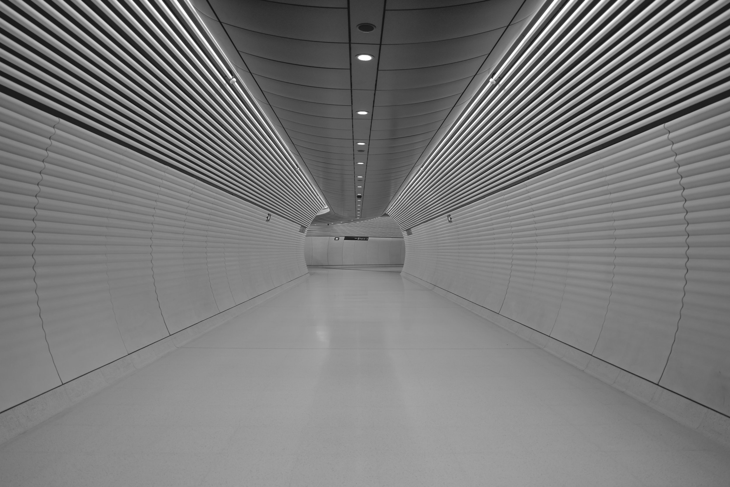 A modern, futuristic tunnel with sleek, curved metallic walls and ceiling. The tunnel is illuminated by evenly spaced overhead lights, creating a clean and minimalist look. The floor is smooth and reflective.