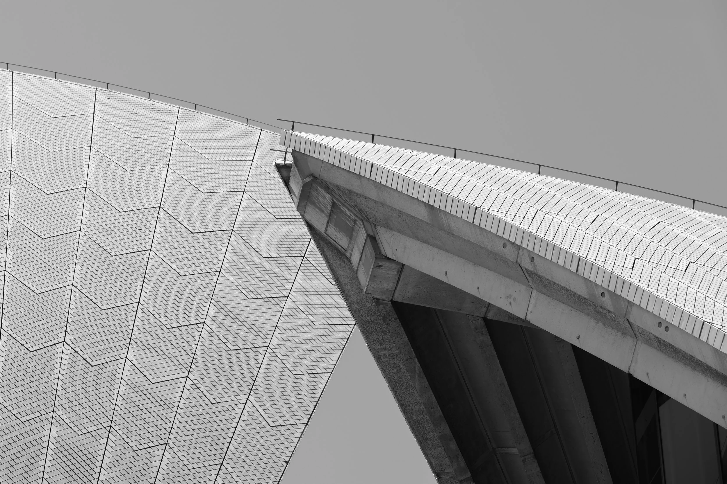 A grayscale photograph of a modern architectural structure with a peaked roof, covered in a grid pattern of tiles or panels, and supported by concrete beams. The background features a clear, gray sky.