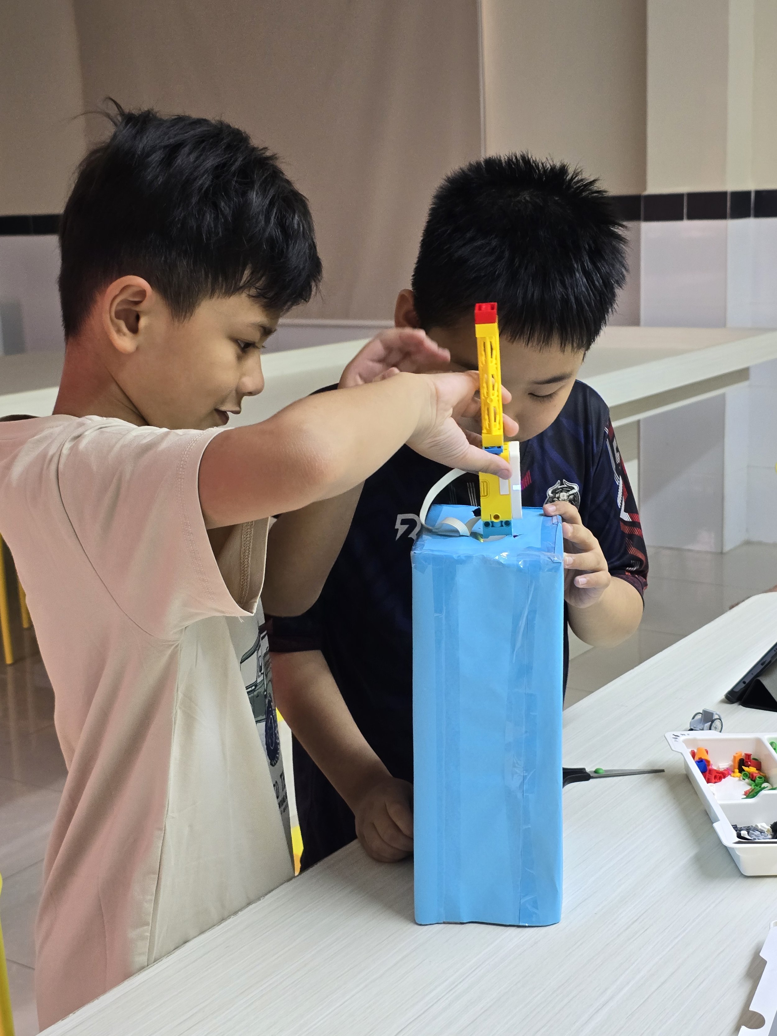Two young boys are building a tall structure with LEGO bricks inside a blue box. They are focused on their activity, with one boy in a beige shirt and the other in a dark shirt. The setting appears to be indoors, possibly in a classroom or play area, with a white table and some LEGO pieces in a white tray nearby.