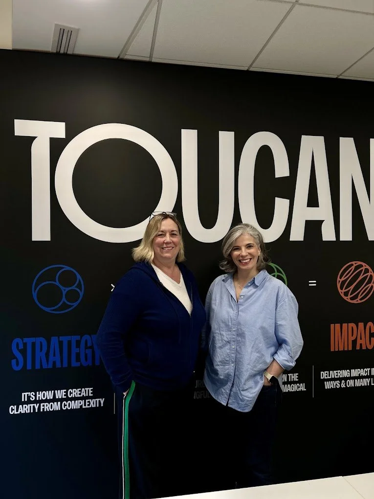 Two women standing in front of a wall with large text that says "TOUCAN" and colorful graphics, smiling for a photo.