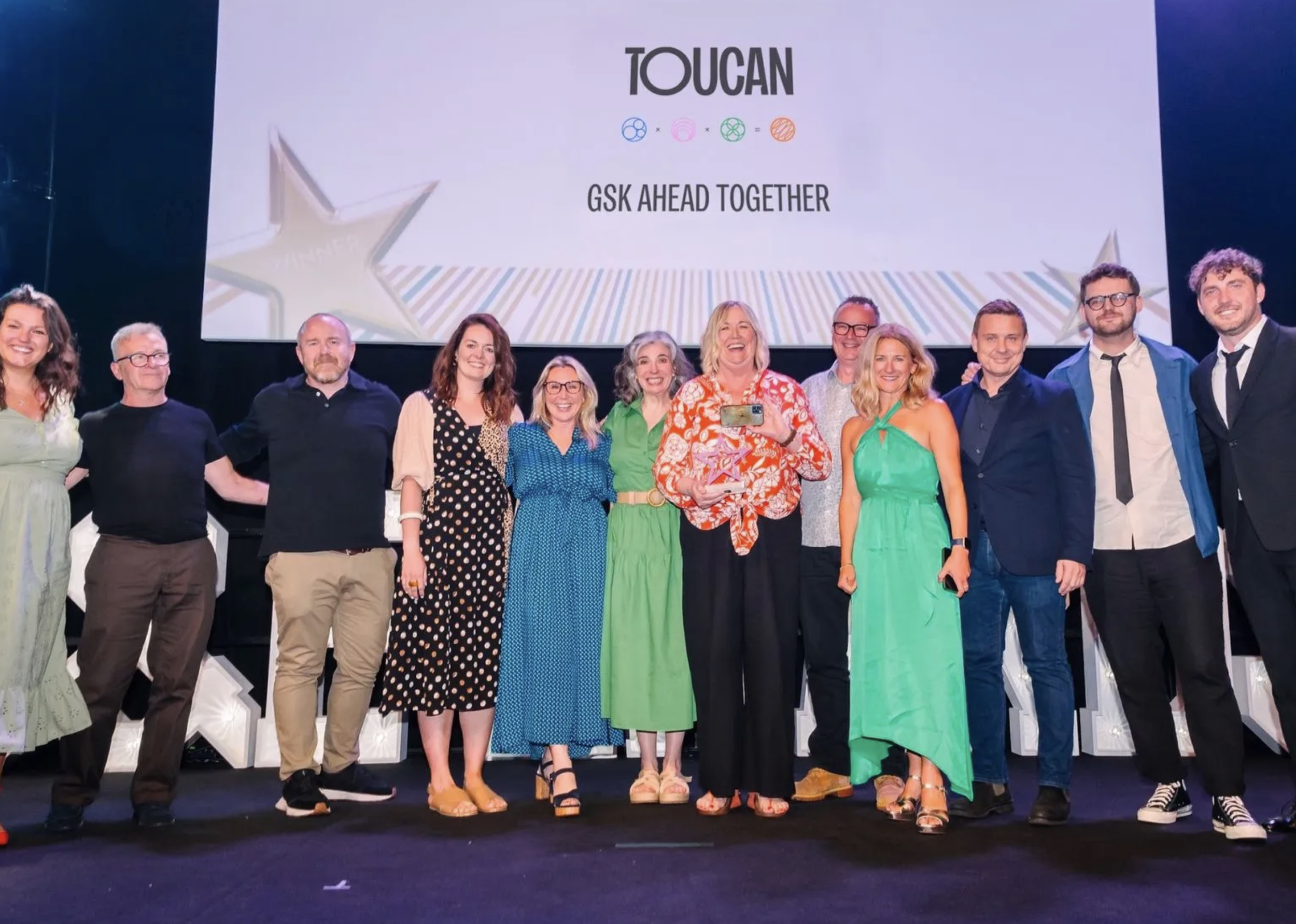 Group of people standing on stage in front of a large screen with the word 'TOUCAN' and the phrase 'GSK AHEAD TOGETHER'. They are smiling, dressed in semi-formal attire, and appear to be celebrating an event or achievement.