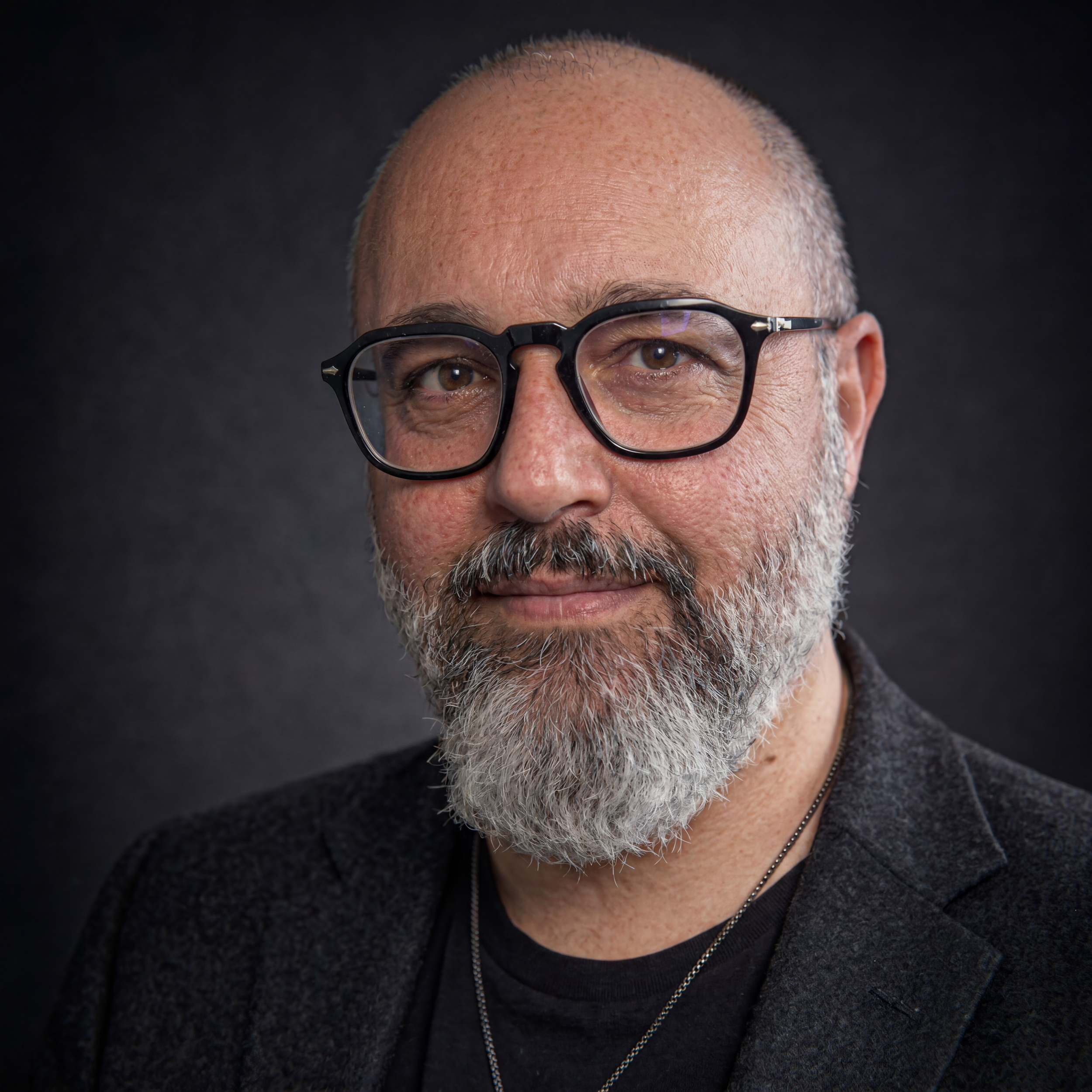 A bald man with glasses and a goatee, smiling, wearing a tan jacket over a black shirt, against a dark gray background.