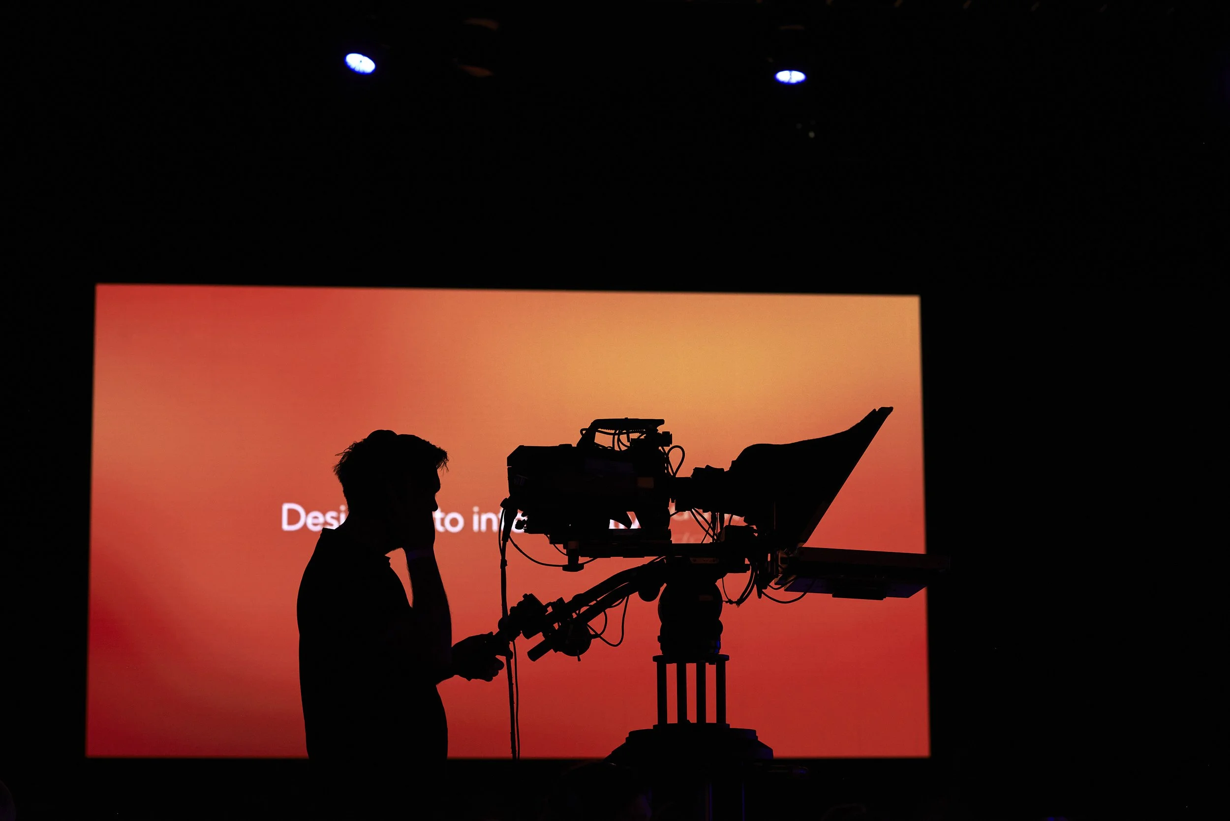 Silhouette of a person operating a camera on a tripod against a large orange gradient screen.