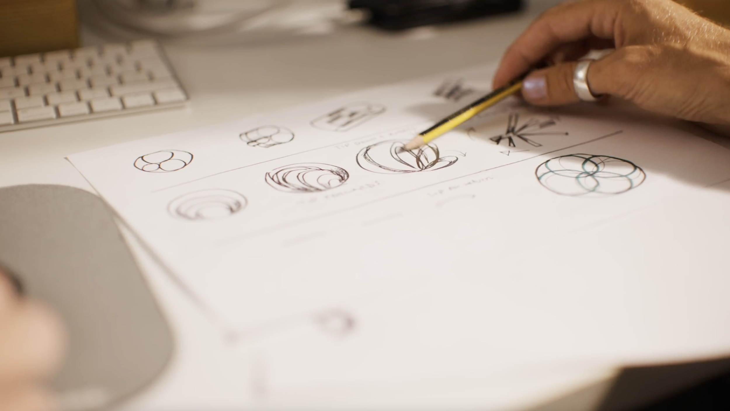 A person is sketching geometric designs on a sheet of paper with a pencil, with various drawings and diagrams visible on the paper.