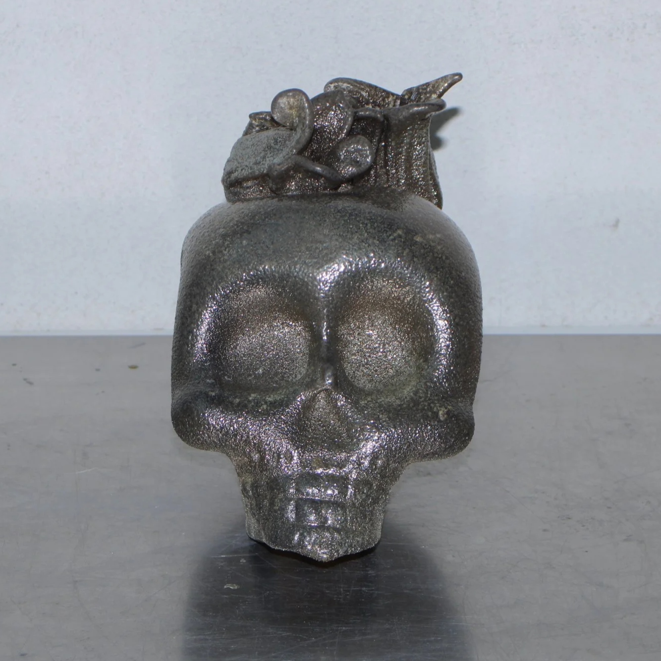 A metallic skull-shaped sculpture with a flower on top, placed on a metallic surface against a plain background.