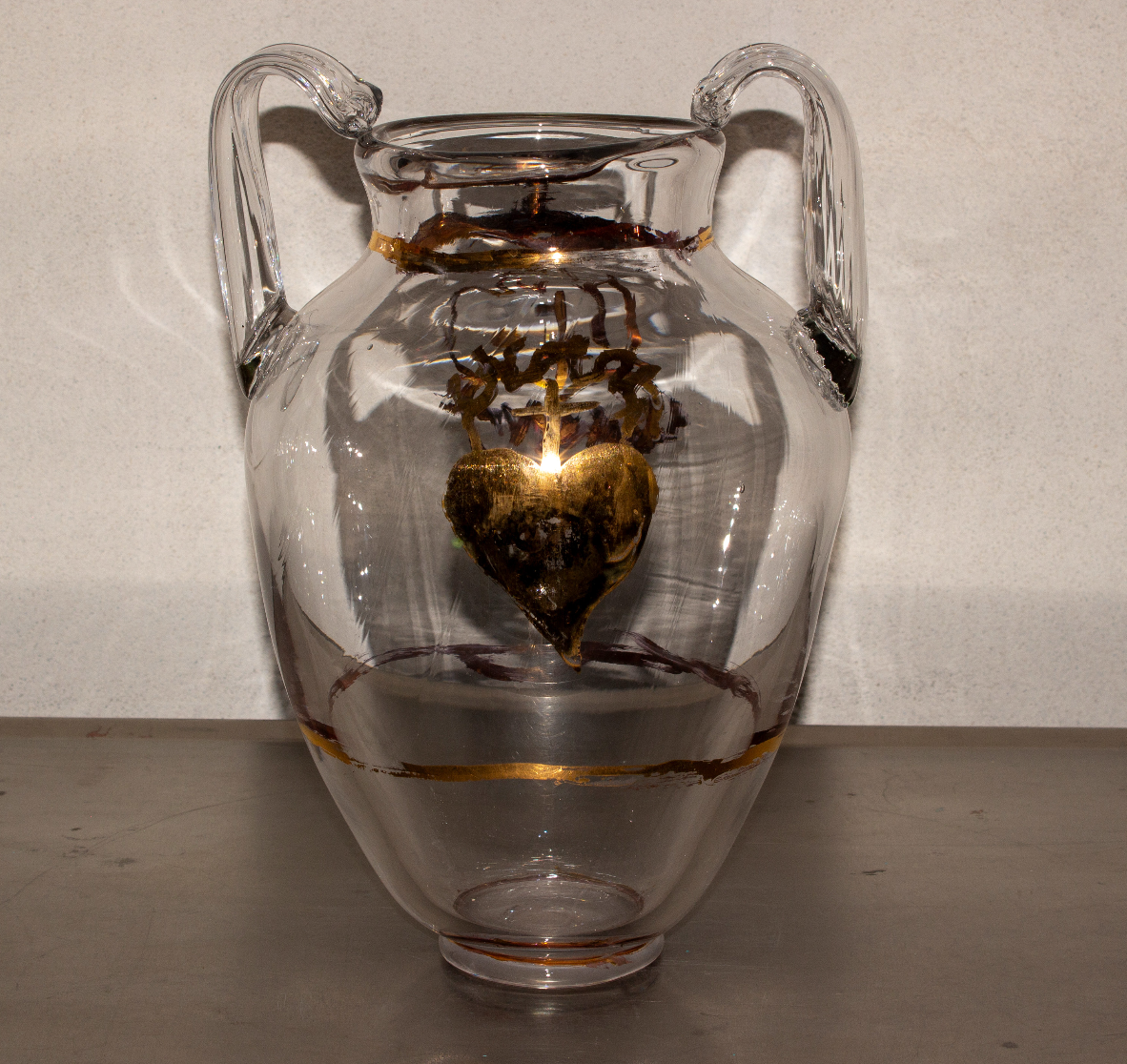 A glass vase with a clear exterior containing a metallic heart-shaped object suspended inside, with a small lamp or light source illuminating the heart.