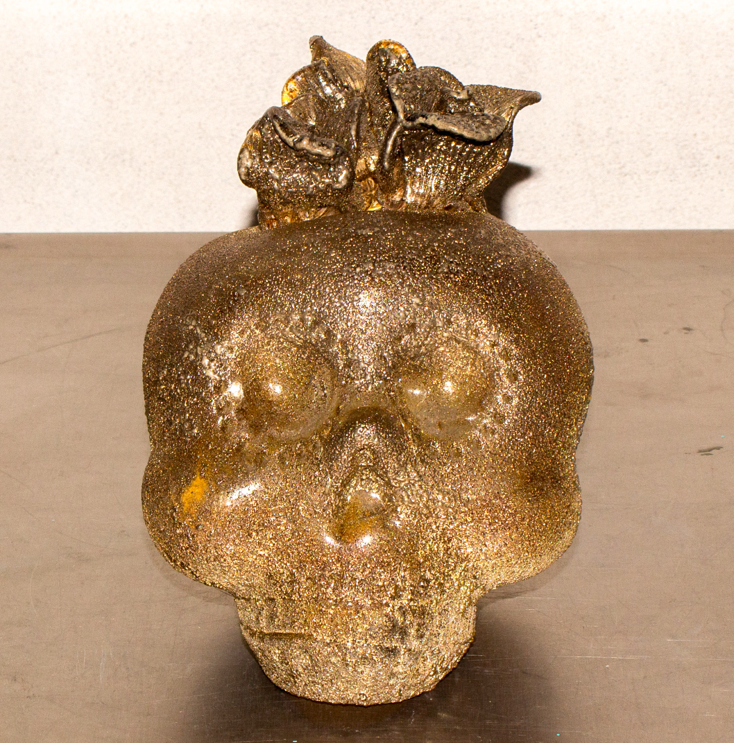 Gold-colored skull sculpture with a decorative top, placed on a flat surface against a plain background.