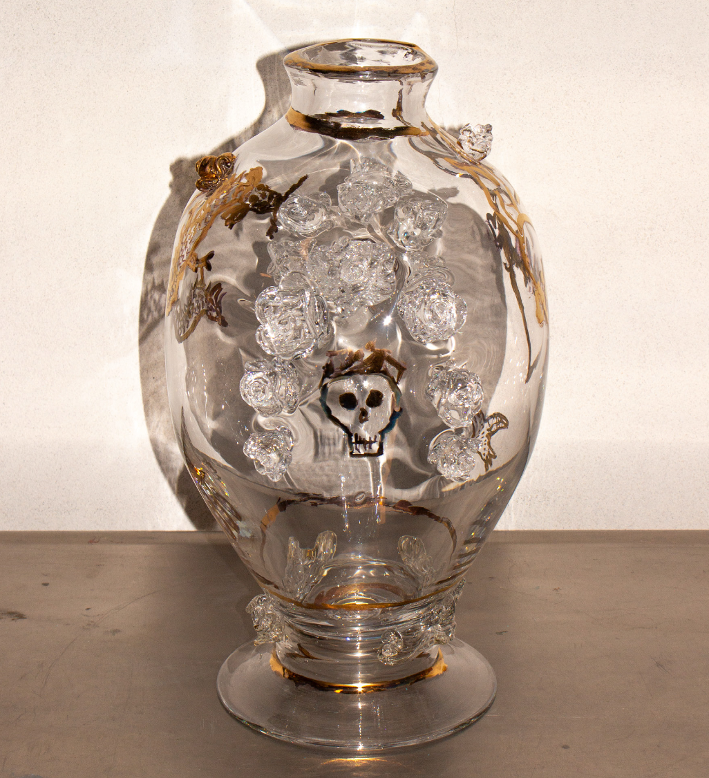 A clear glass vase decorated with gold accents, containing a skull-shaped object surrounded by clear glass roses, with a shadow cast on a white wall behind it.