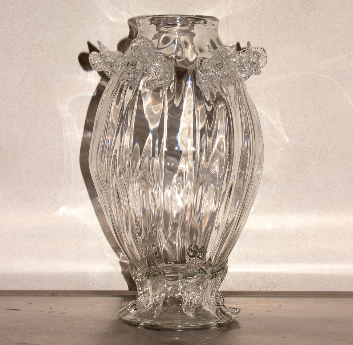 A tall, clear, decorative glass vase with ornate, ruffled edges and intricate patterns, sitting on a light-colored surface with shadows cast behind it.