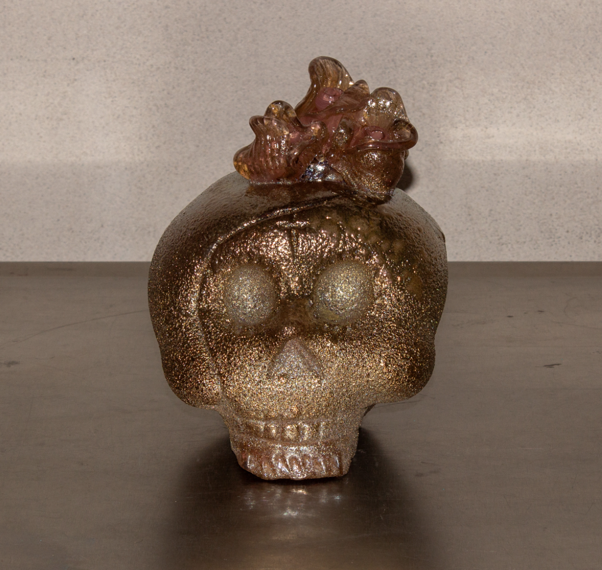 A skull-shaped sculpture covered in gold glitter with a glass or resin top resembling flowers or abstract forms.