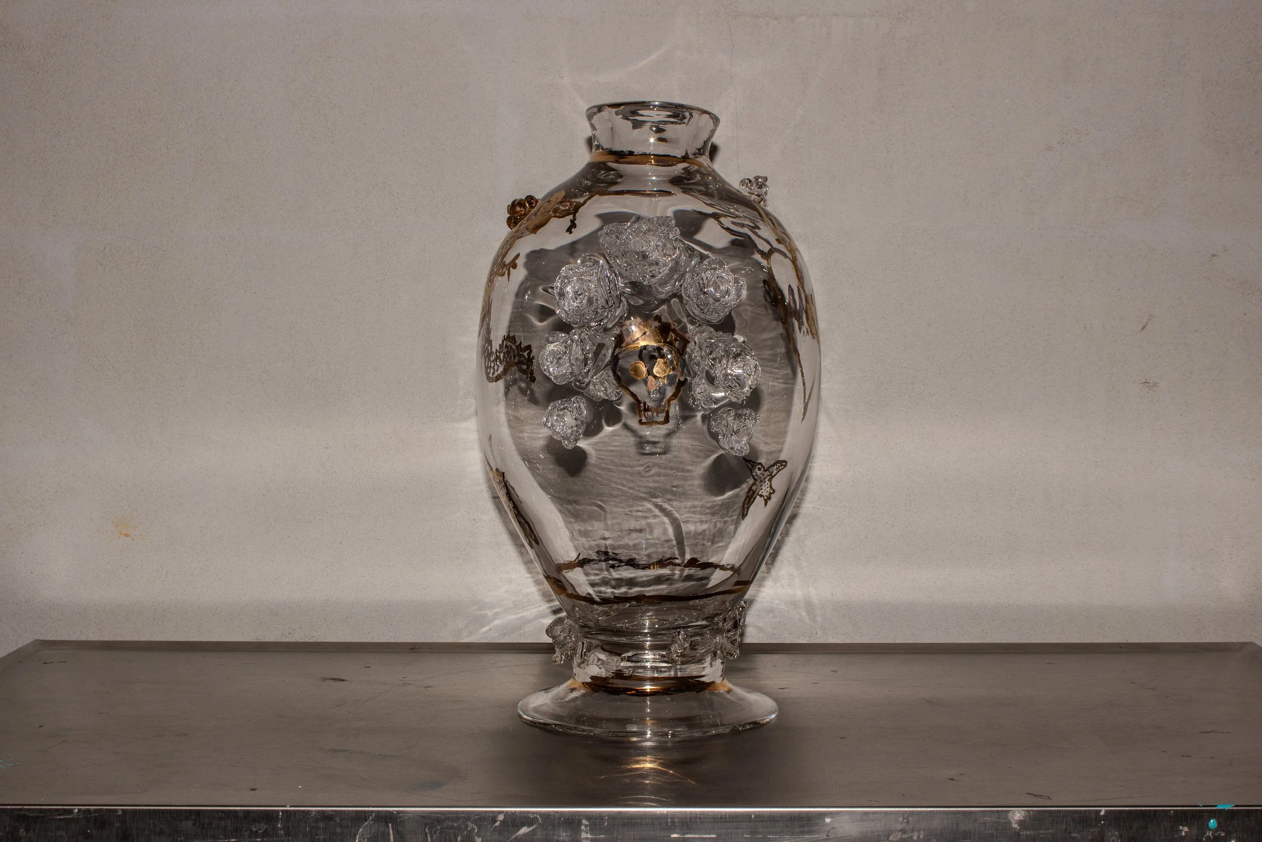 Glass vase with gold accents and decorative glass flowers, butterflies, and a skull, placed on a reflective surface against a plain background.
