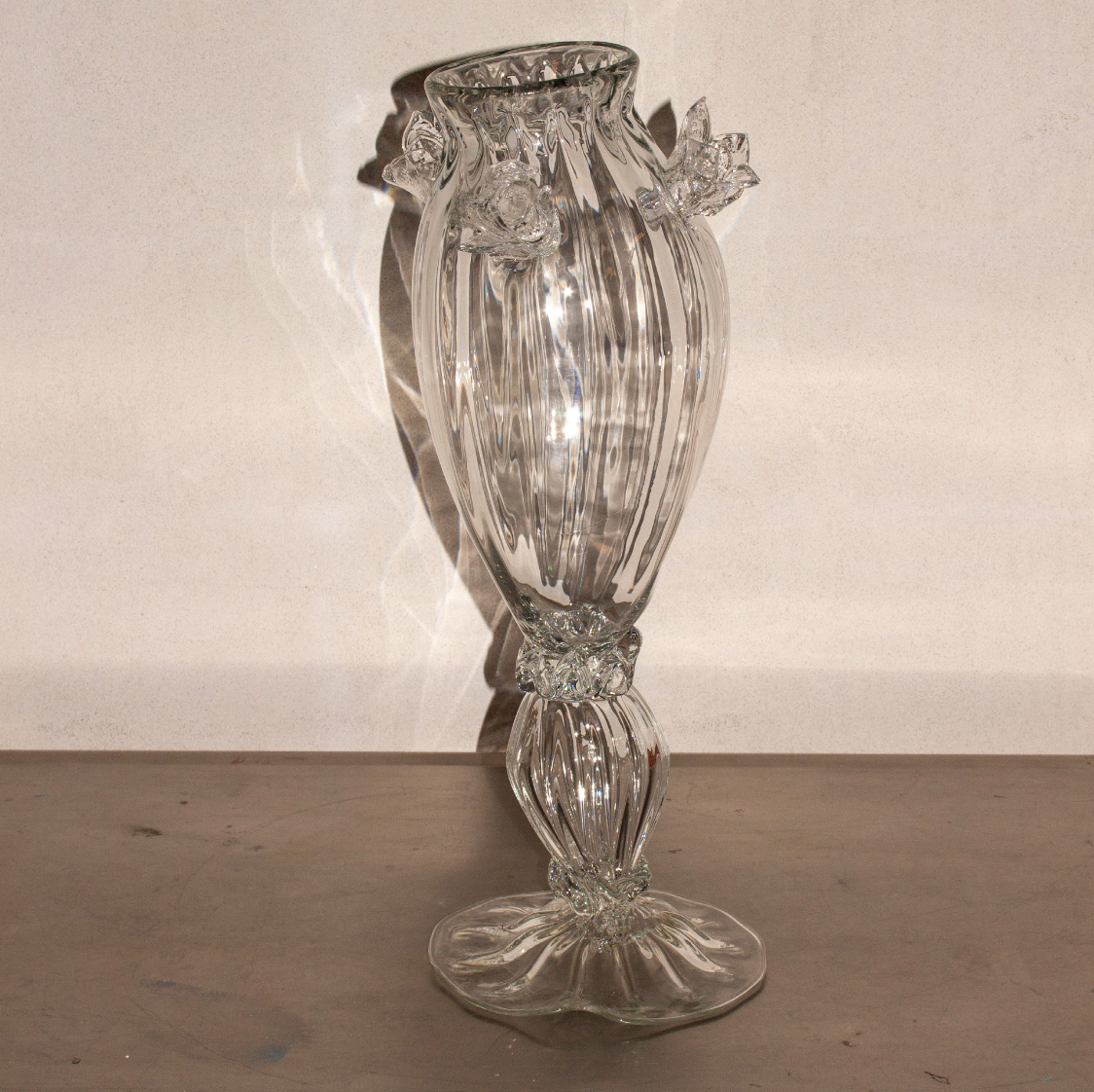 A tall, clear glass vase with a fluted body and ornate, decorative accents near the top and bottom, placed on a wooden surface with a plain background.