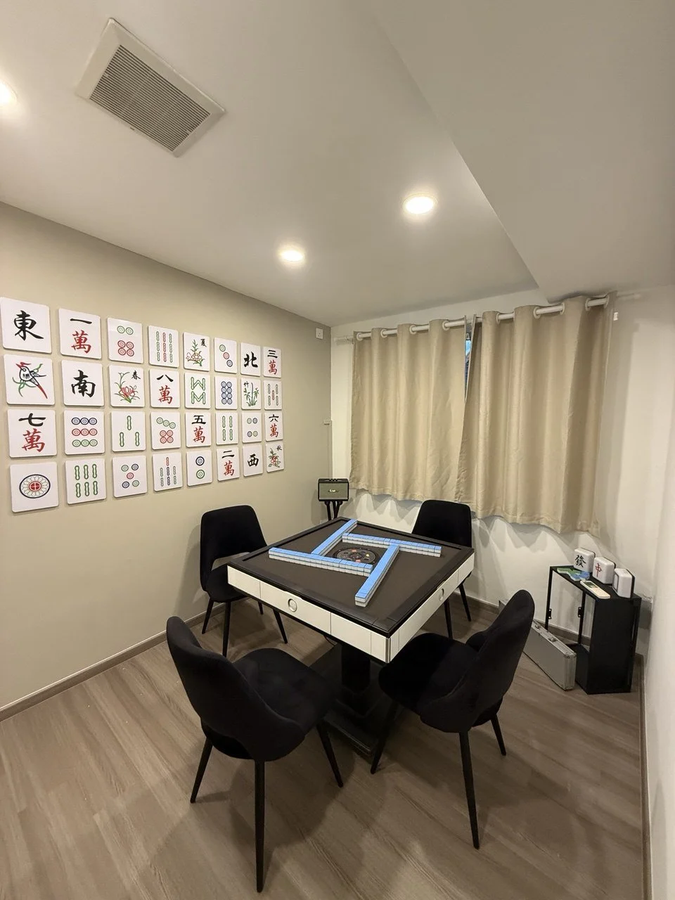 A Mah Jong game room with a square Mah Jong table surrounded by four black chairs. The wall features Mah Jong tiles on display, and there are beige curtains covering a window. The room has a light-colored wood floor and minimal décor.