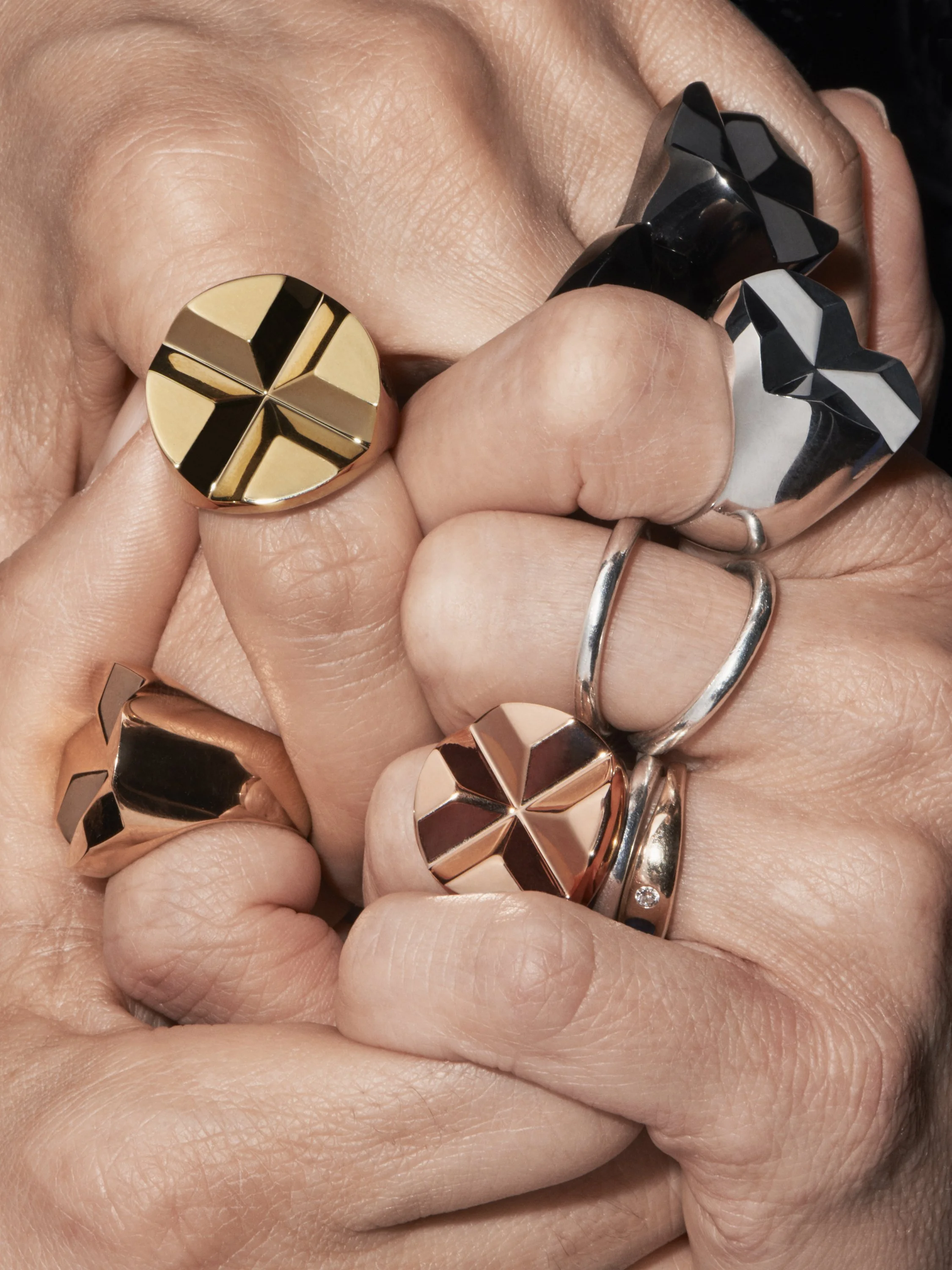 Close-up of hands wearing multiple large geometric rings in gold, silver, black, and rose gold colors.