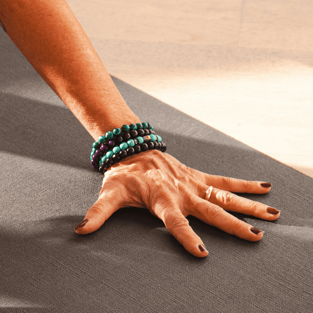 Close-up of a person's hand and wrist with multiple beaded bracelets, pressing down on a yoga mat during an asana.