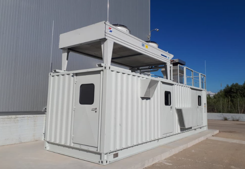 White industrial modular container unit with rooftop equipment, doors, vents, and safety railing outside a warehouse