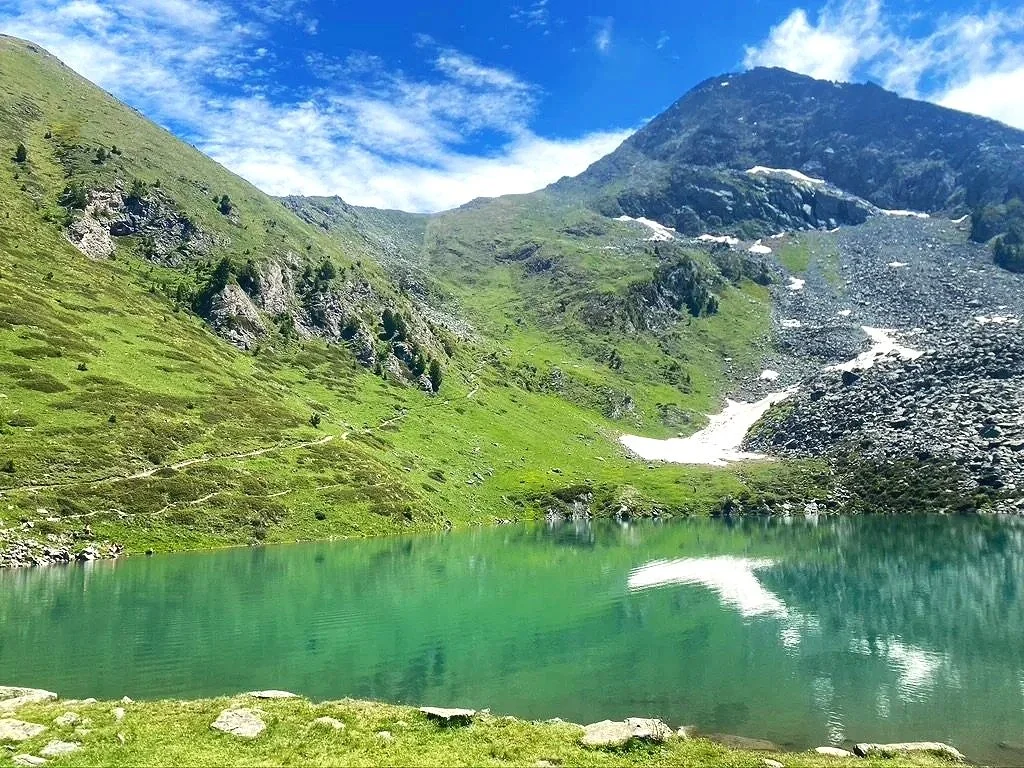 Aosta Valley Italy - mountain lake with turquoise water surrounded by green hills and rocky slopes, with a snow patch, under a partly cloudy sky.