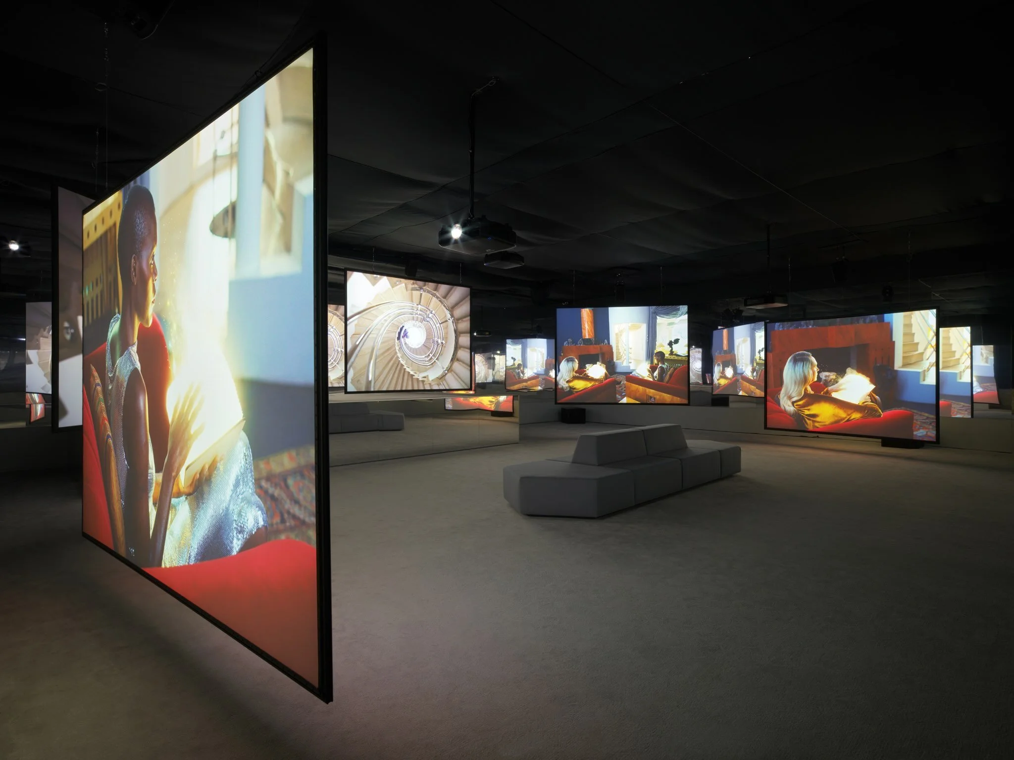 Room with multiple large LCD screens showing different images, dark ceiling with hanging projectors, gray couches, and people sitting and talking