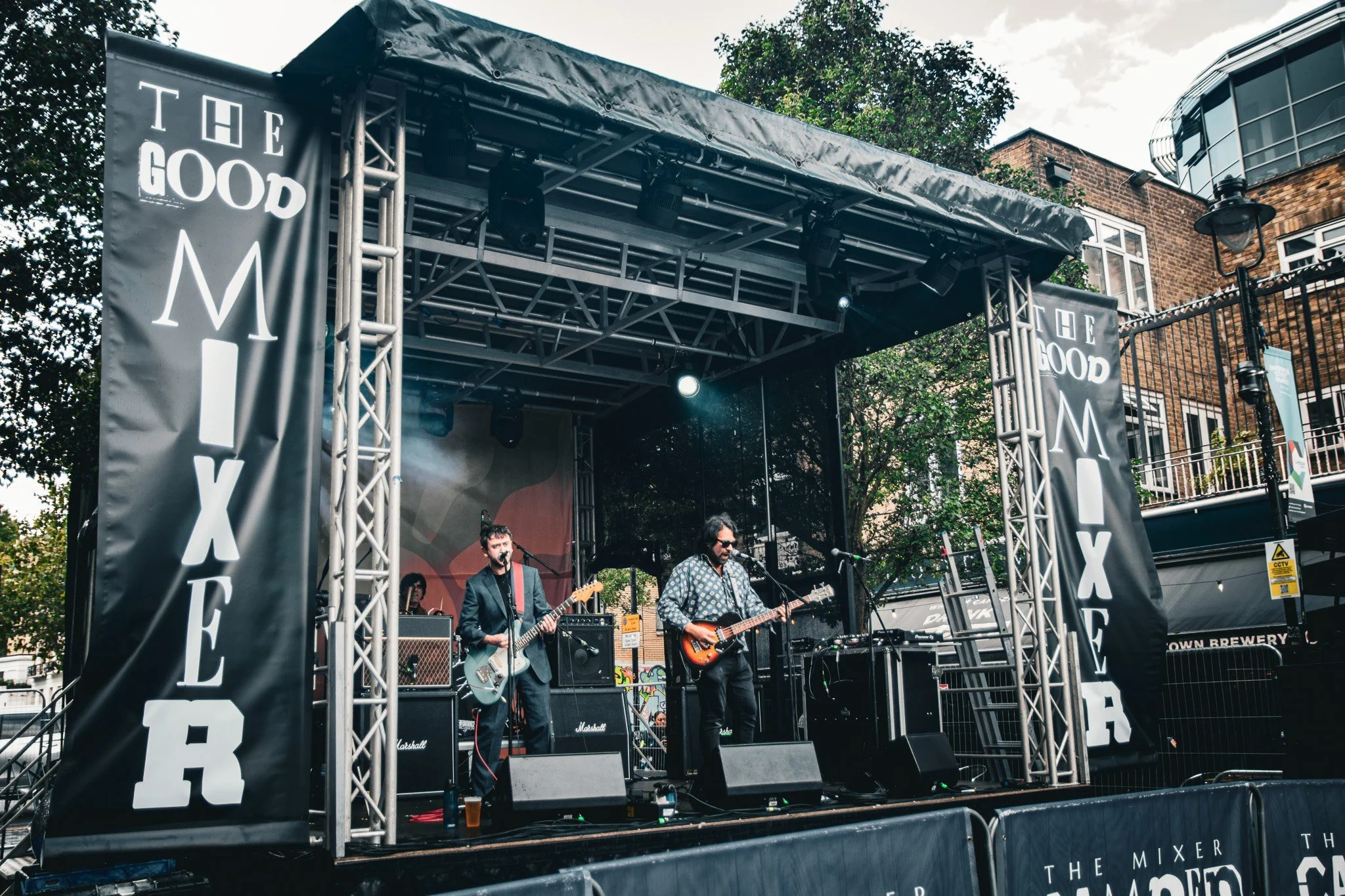 A live outdoor band performance on a stage with musical instruments and amplifiers, with banners displaying "THE GOOD MEXICAN" at the sides of the stage, and a brick building with trees and a cloudy sky in the background.