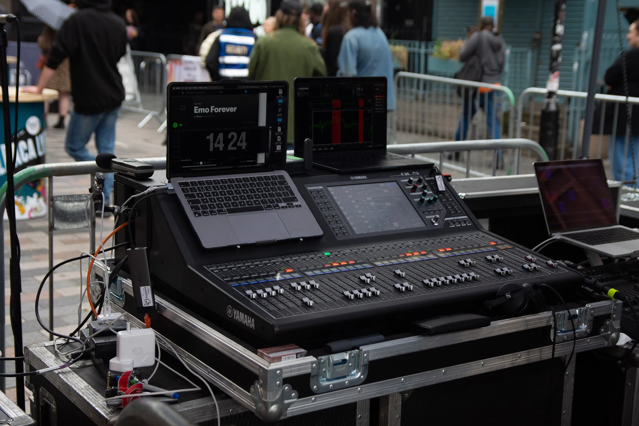 Sound equipment including a mixer, laptops, and audio controls at an outdoor event with people in the background.