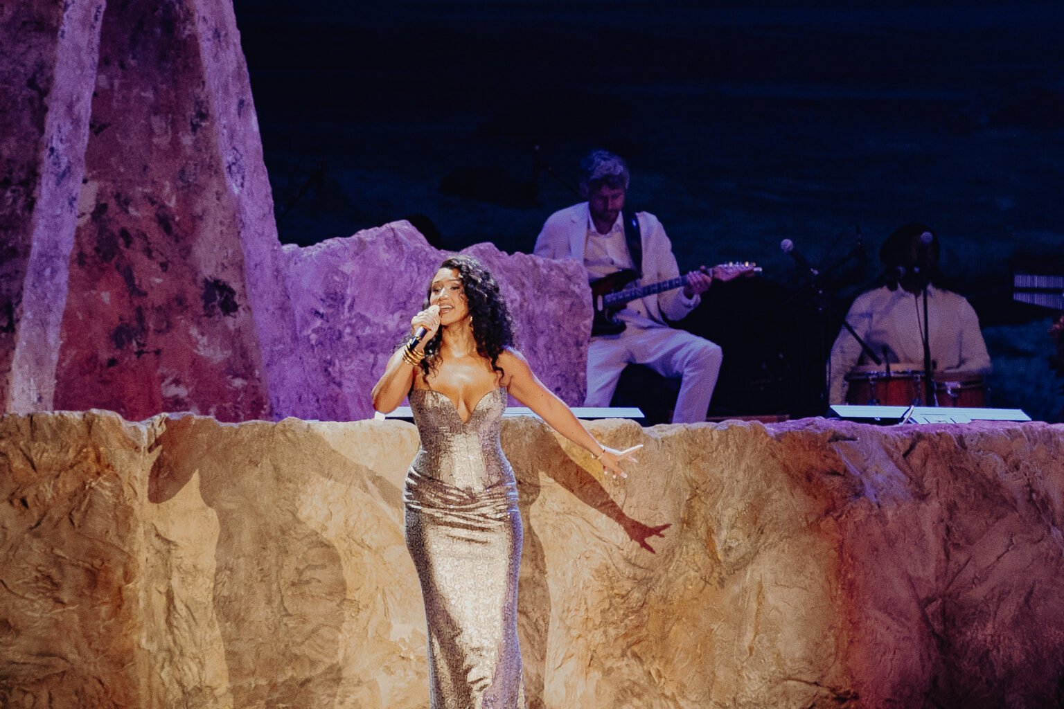 Cleo Sol singing on stage with a microphone, wearing a shiny gown, with a band playing instruments behind her, set against a rocky, desert-like backdrop.