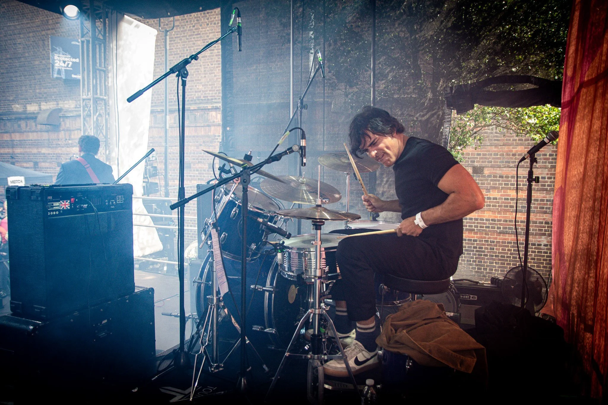 A male musician playing drums on stage during a live performance, with various microphones and equipment present, and another musician in the background near a keyboard.