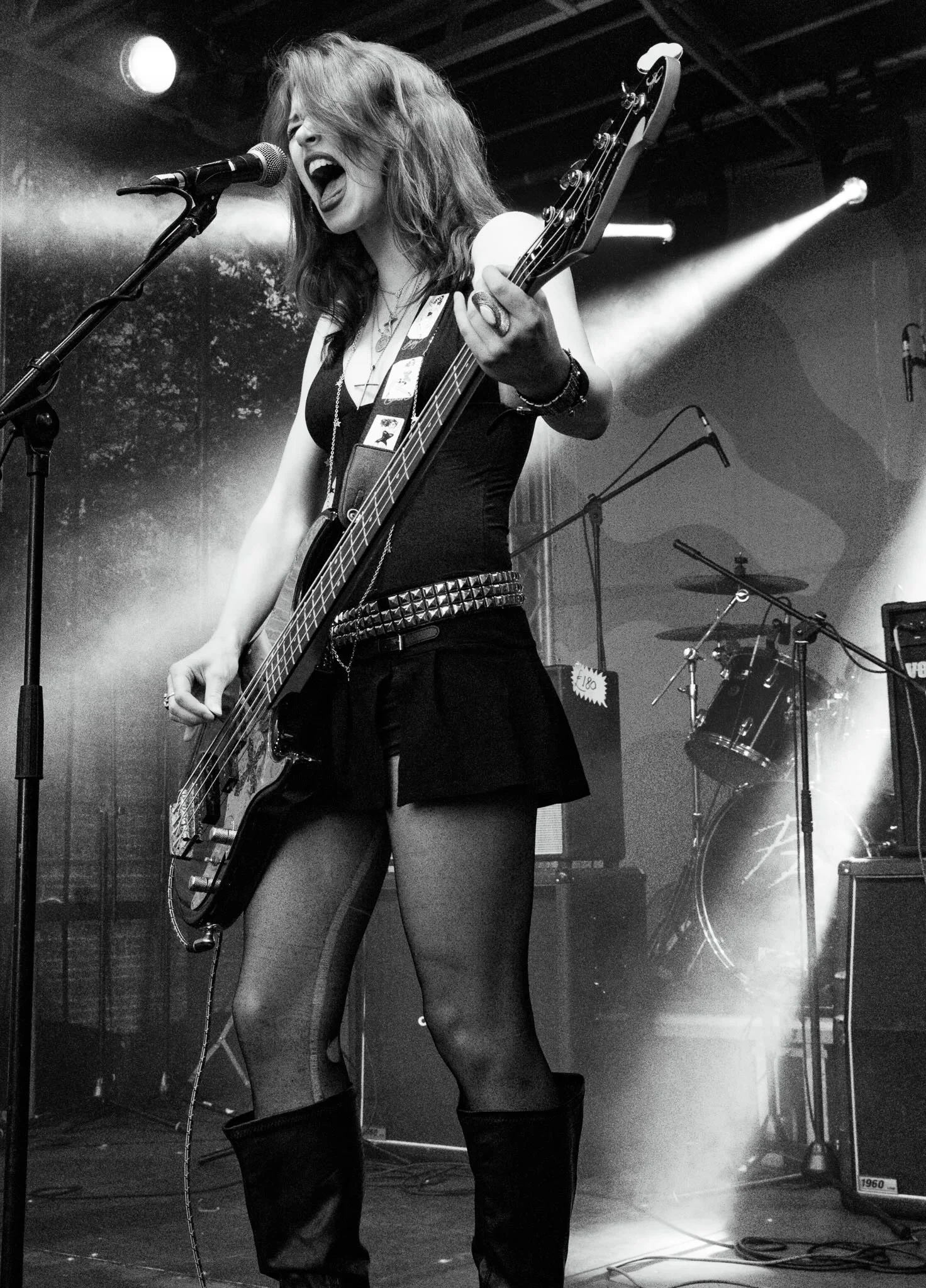A female musician passionately singing into a microphone while playing an electric bass guitar on stage, with stage lighting and musical equipment visible in the background.