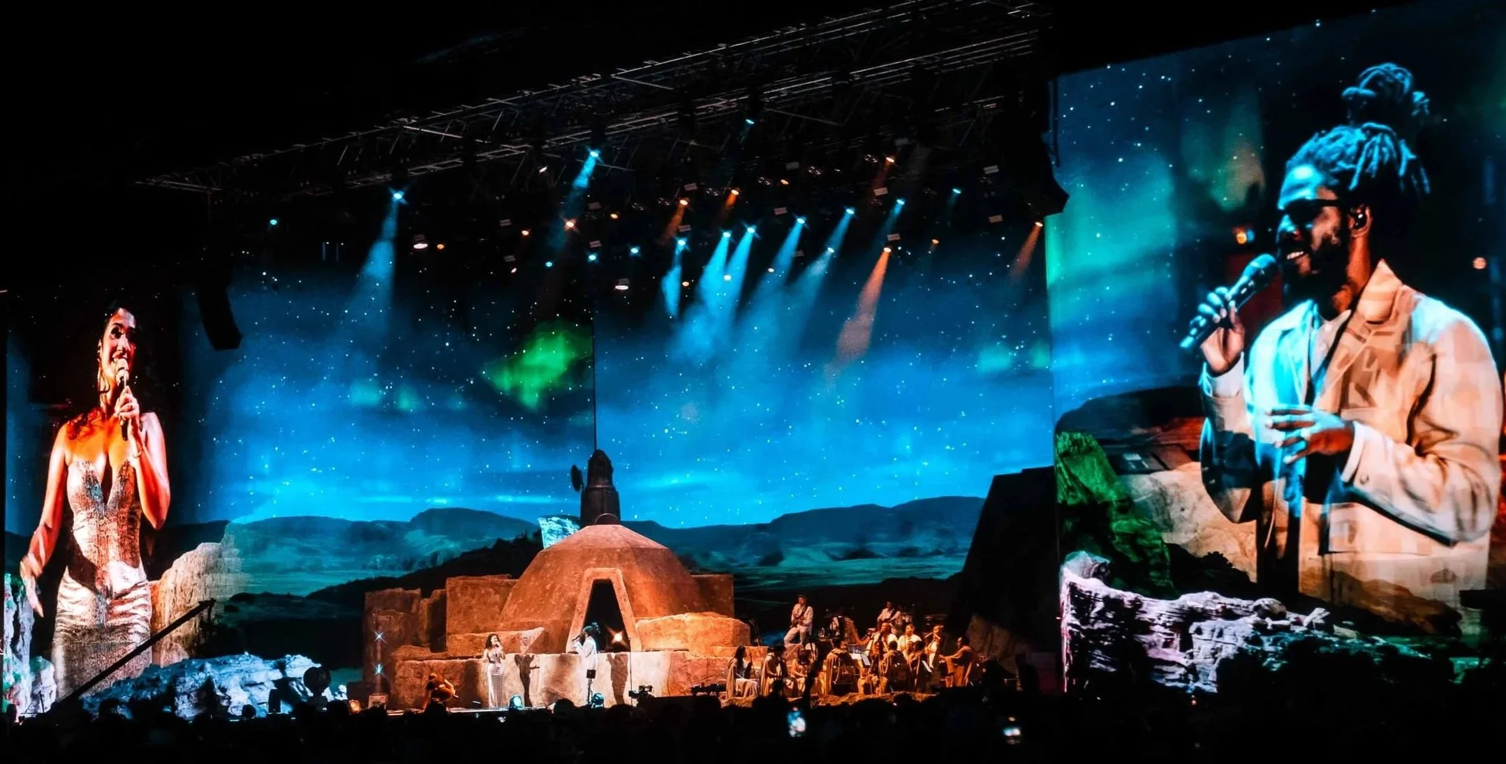 A stage with two large screens showing close-ups of performers: one woman on the left in a dress, and one woman on the right with glasses and dreadlocks holding a microphone. The backdrop depicts a starry night sky and a landscape resembling the moon or Mars with rocks and hills. Stage lighting includes blue and orange spotlights.