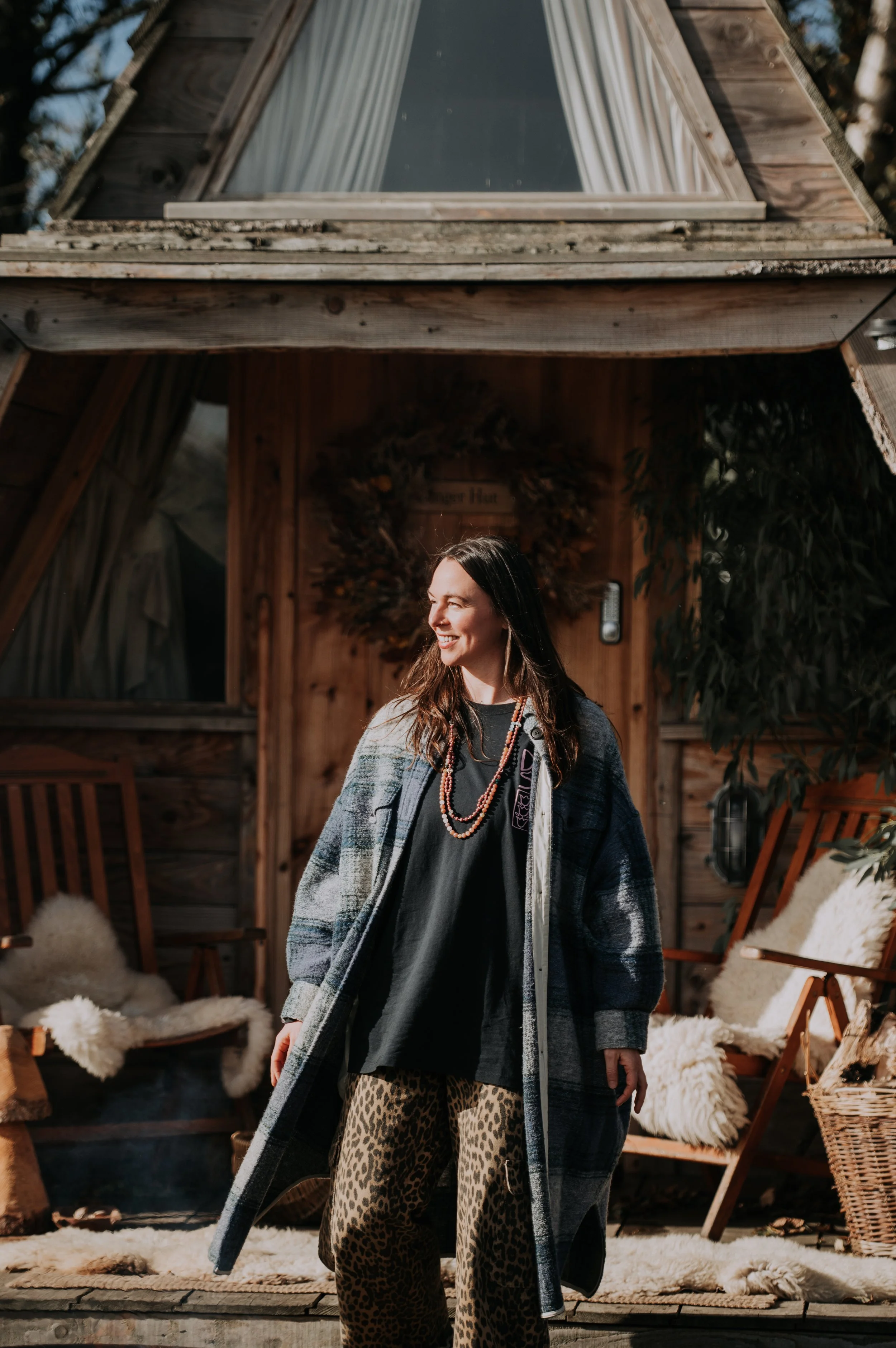 Woman standing outdoors in front of a wooden cabin, wearing leopard-print pants, a black t-shirt, and a striped coat, smiling and looking to the side.