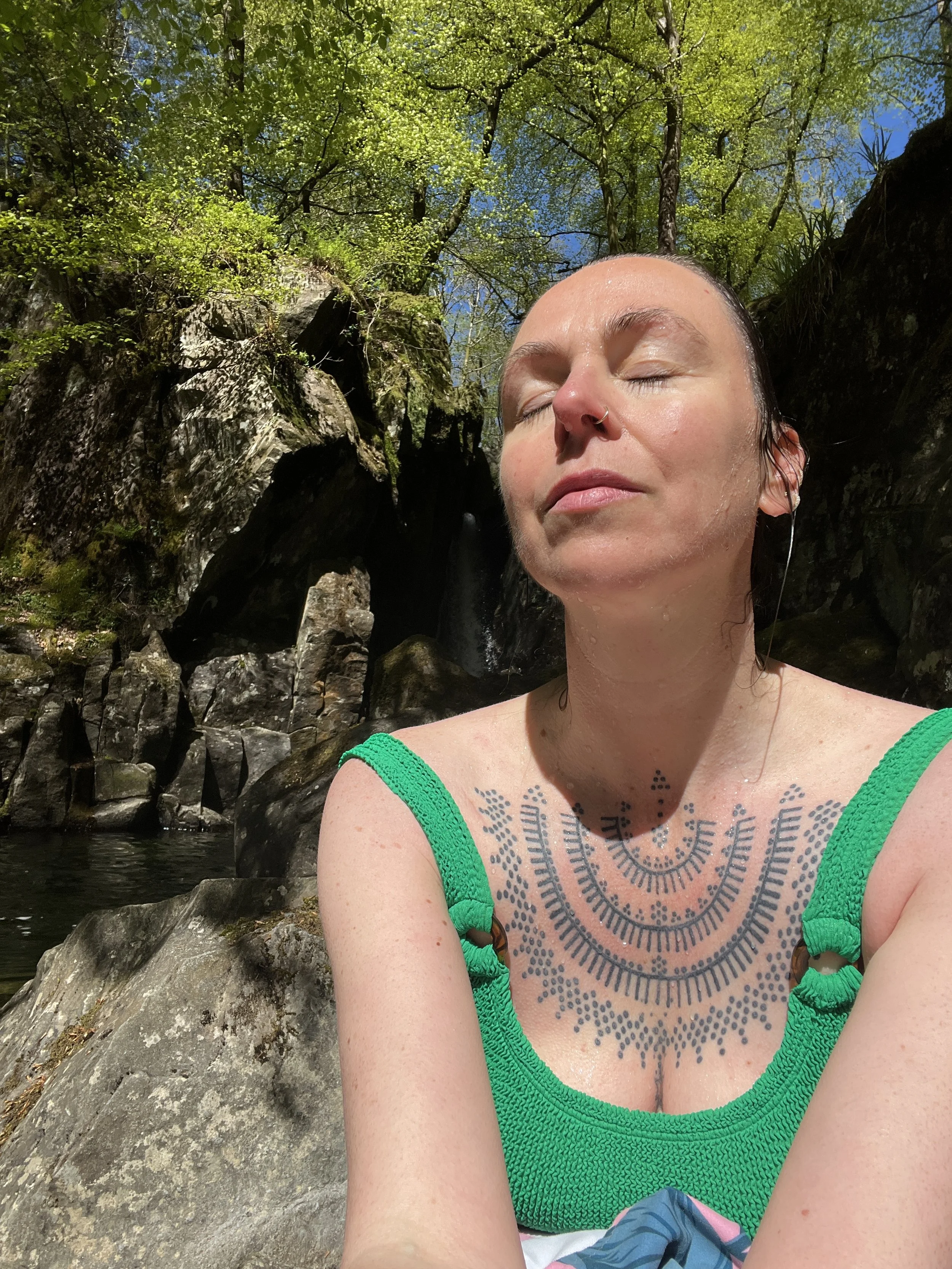 A woman with closed eyes, tattoos on her chest, and a nose ring, sitting among rocks and trees with green foliage and a blue sky in the background.