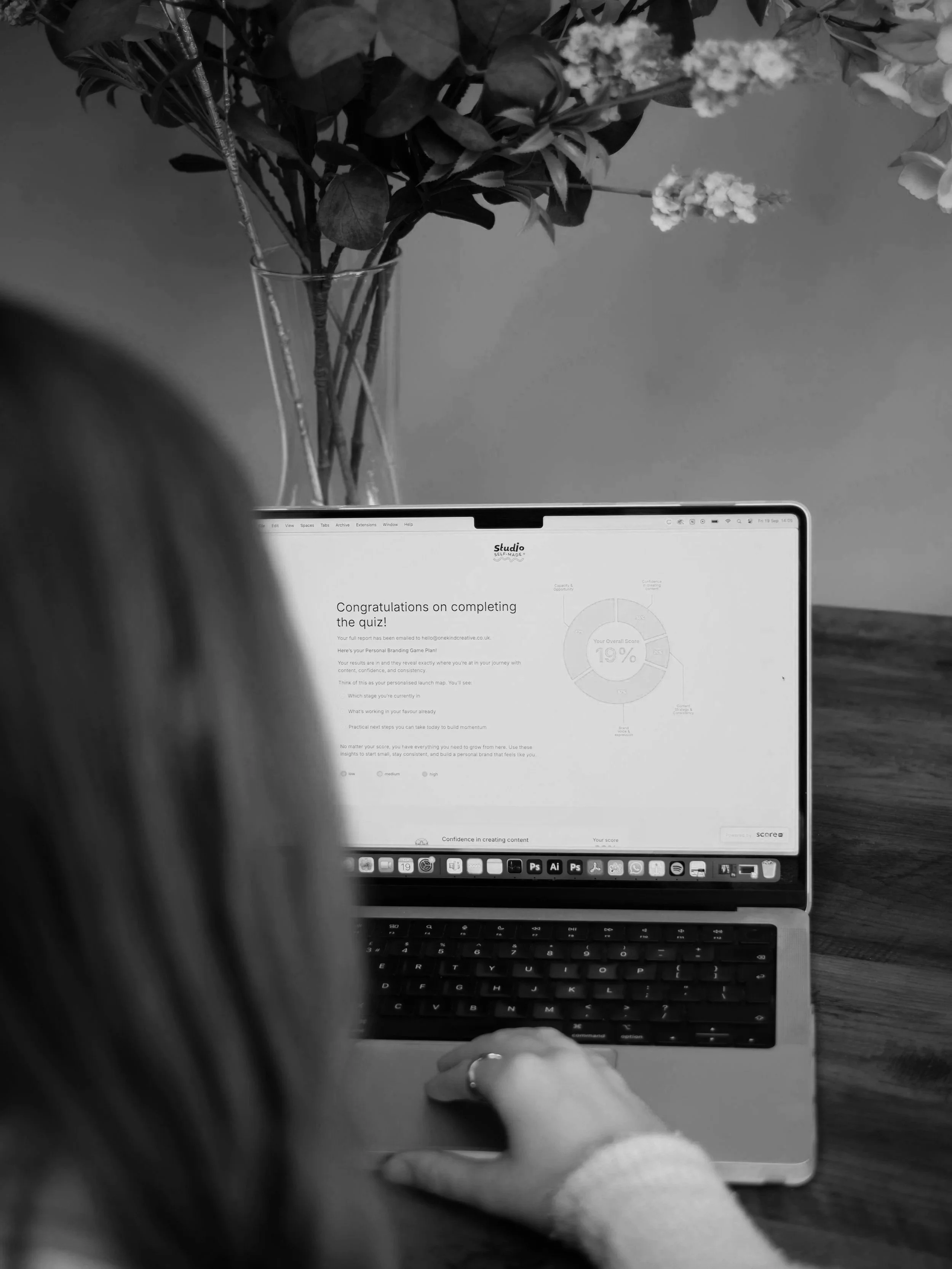 A person using a laptop at a wooden table with a vase of flowers in the background. The laptop screen displays a congratulatory message for completing a quiz.